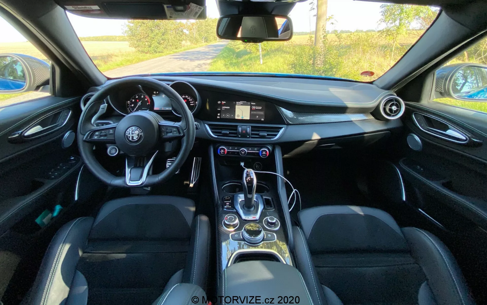 Vista del interior de un Alfa Romeo Giulia 2016 desde la perspectiva del conductor mostrando un volante de fondo plano con múltiples botones de control, un sistema de infoentretenimiento digital en el tablero, un reloj analógico, ventiladores circulares, una elegante palanca de cambios, selector de modos de conducción, portavasos, ventanas eléctricas, asientos de cuero con refuerzos laterales y características tecnológicas modernas en general.