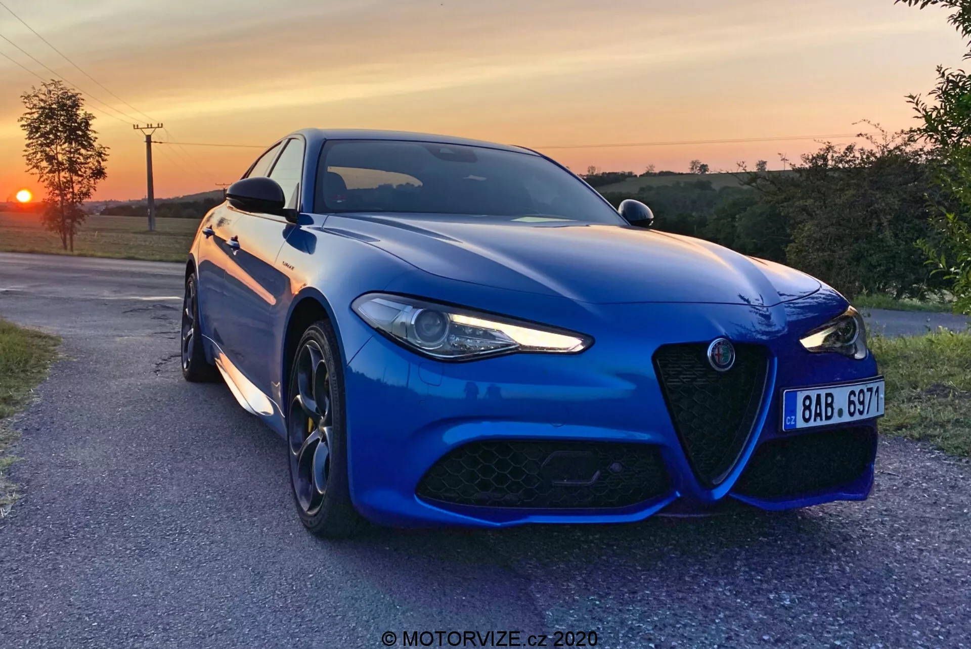Un sedán deportivo Alfa-Romeo Giulia 2016 azul capturado por la tarde con una puesta de sol en el fondo, posicionado ligeramente en ángulo hacia la cámara en un camino pavimentado del campo, mostrando su distintiva parrilla delantera, diseño agresivo de faros LED, grandes ruedas de aleación de múltiples radios y un sistema de frenos de alto rendimiento. La pintura metálica, el parachoques delantero aerodinámico, las líneas esculpidas del capó y los sensores avanzados de asistencia a la conducción destacan la estética deportiva y la tecnología moderna del coche. Hay una marca de agua 'MOTORVIZE.cz 2020' presente.
