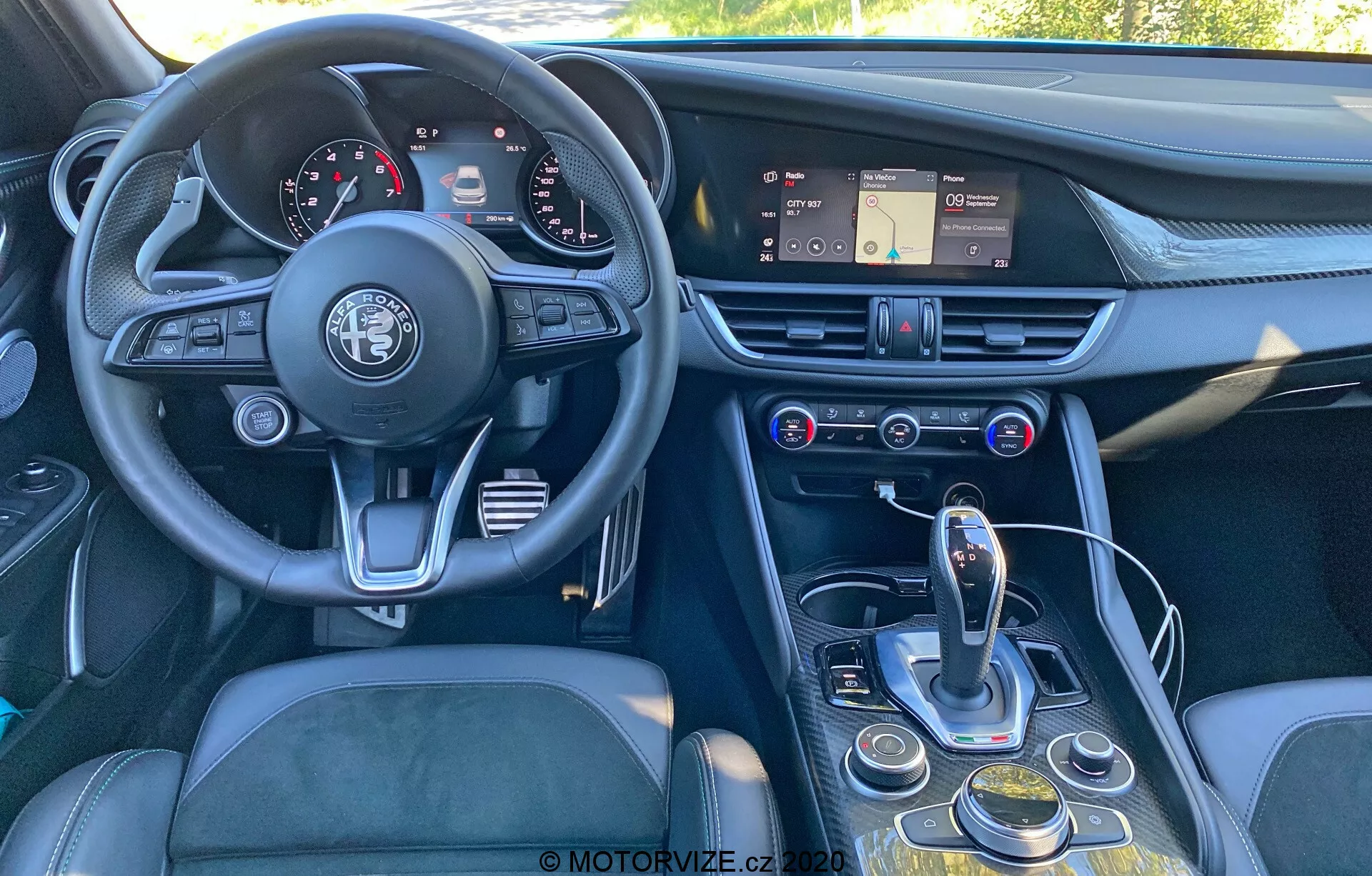 Vista interior de un Alfa Romeo Giulia 2016 desde un ángulo levemente descentrado, de atrás hacia adelante, mostrando el tablero, el volante, la consola central y parte del asiento del conductor. La imagen destaca el volante de tres radios con el emblema de Alfa Romeo, un panel de instrumentos con diales analógicos y pantallas digitales, un tablero con un sistema de infoentretenimiento con pantalla táctil, ventilas de aire y controles de clima y multimedia. La consola central incluye un selector de transmisión automática, un dial giratorio, botones para la navegación de infoentretenimiento y configuraciones del vehículo, y un posible selector de modos. El diseño enfatiza la ergonomía y accesibilidad, con materiales como cuero, metal cepillado y plástico de alta calidad para una sensación de lujo.