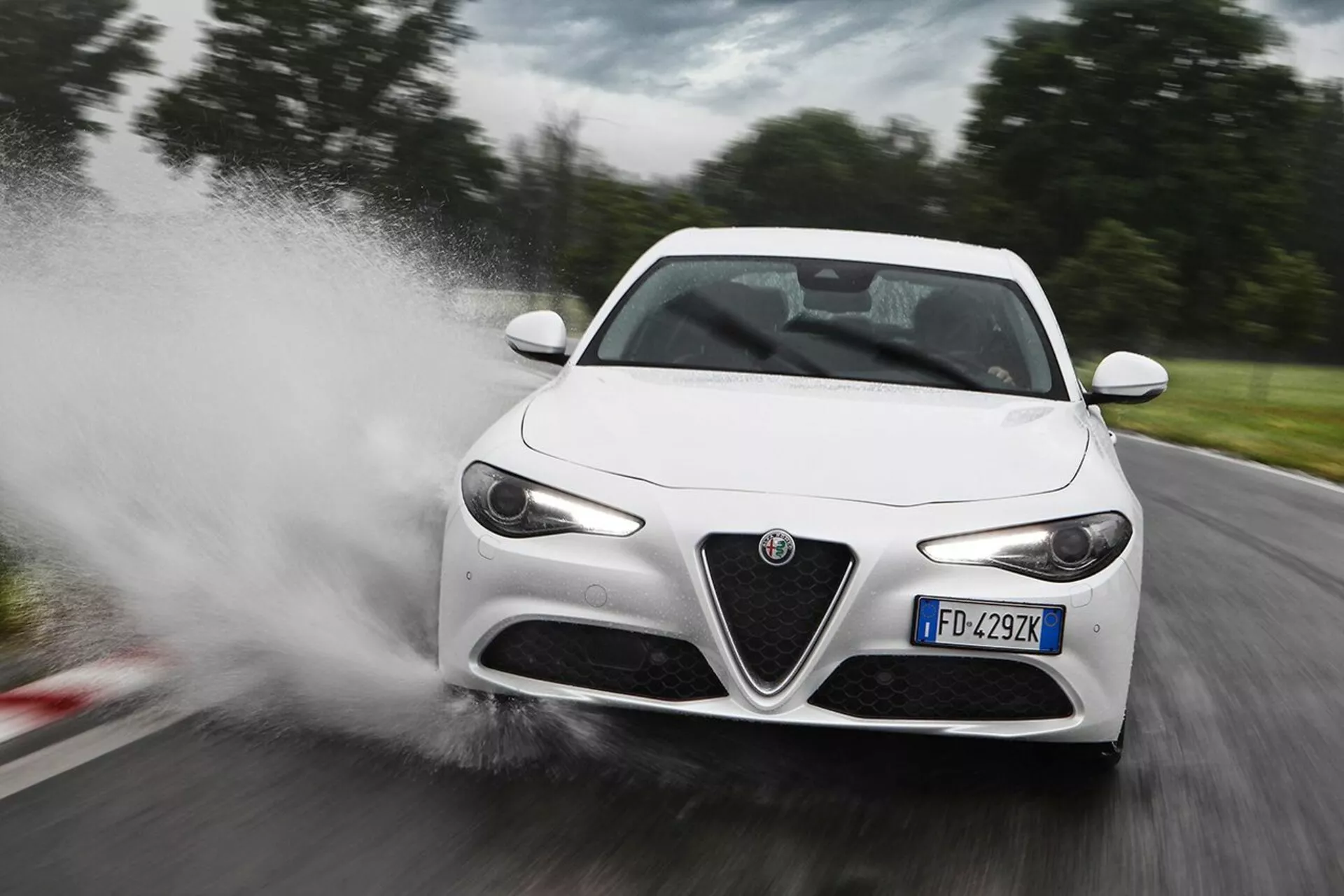 Deportivo Alfa Romeo Giulia blanco de 2016 acelerando a través del agua, causando un chapoteo, en una vista frontal de tres cuartos destacando sus faros LED, parrilla frontal distintiva, espejos laterales elegantes y líneas aerodinámicas