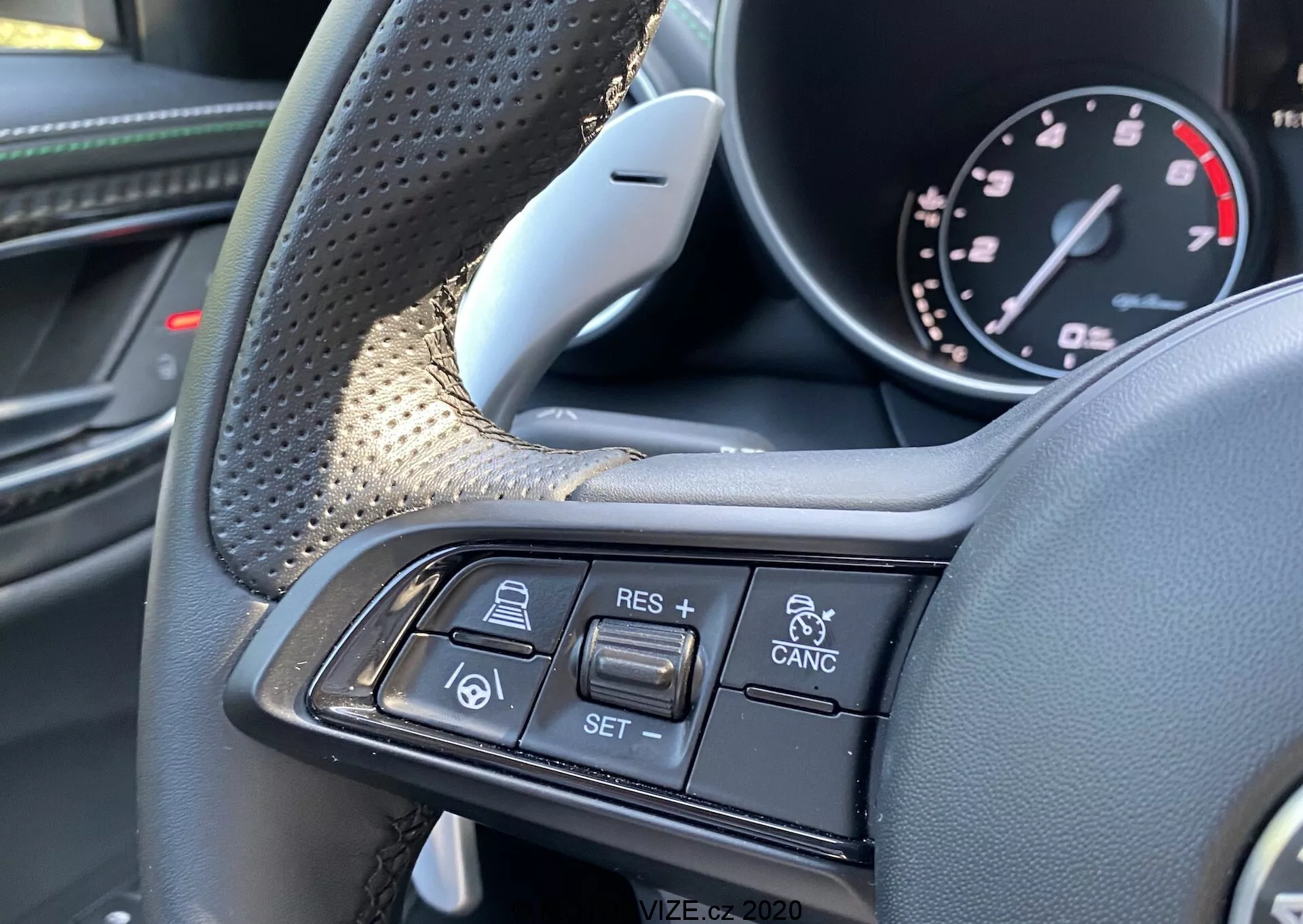 Vista de primer plano del interior del Alfa-Romeo Giulia 2016 que muestra el volante y sus controles multifunción desde la perspectiva del conductor. Se pueden ver las áreas de agarre texturizadas y materiales lujosos como cuero y posiblemente fibra de carbono en el lado izquierdo. La rama derecha presenta botones para el control de crucero y la tecnología de crucero adaptativo, incluidos + RES, - SET, CANCEL, un icono de coche siguiendo a coche y alertas de colisión de vehículos. La imagen se centra exclusivamente en estos controles, destacando el diseño premium y las características avanzadas de asistencia al conductor.