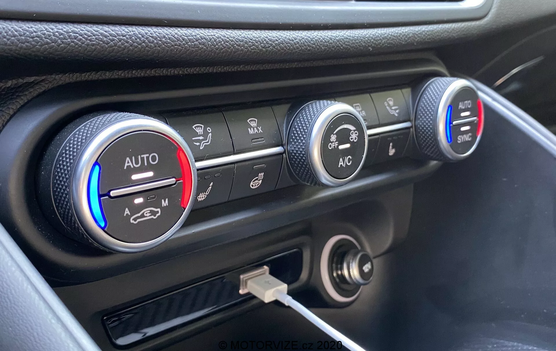 Primer plano del sistema de control climático en el tablero del Alfa Romeo Giulia 2016, con tres perillas circulares. La perilla izquierda indica configuraciones de temperatura con bandas azules y rojas y una configuración 'AUTO', parcialmente iluminada en rojo. La perilla del medio muestra configuraciones de velocidad del ventilador, resaltadas a una velocidad más baja. La perilla derecha muestra varios modos de distribución de aire con botones 'AC' y 'SYNC'. Debajo de las perillas hay una toma de corriente de 12 voltios y un puerto USB. El tablero tiene un acabado en negro con un acento plateado alrededor de los controles de clima, capturado desde un ángulo elevado.