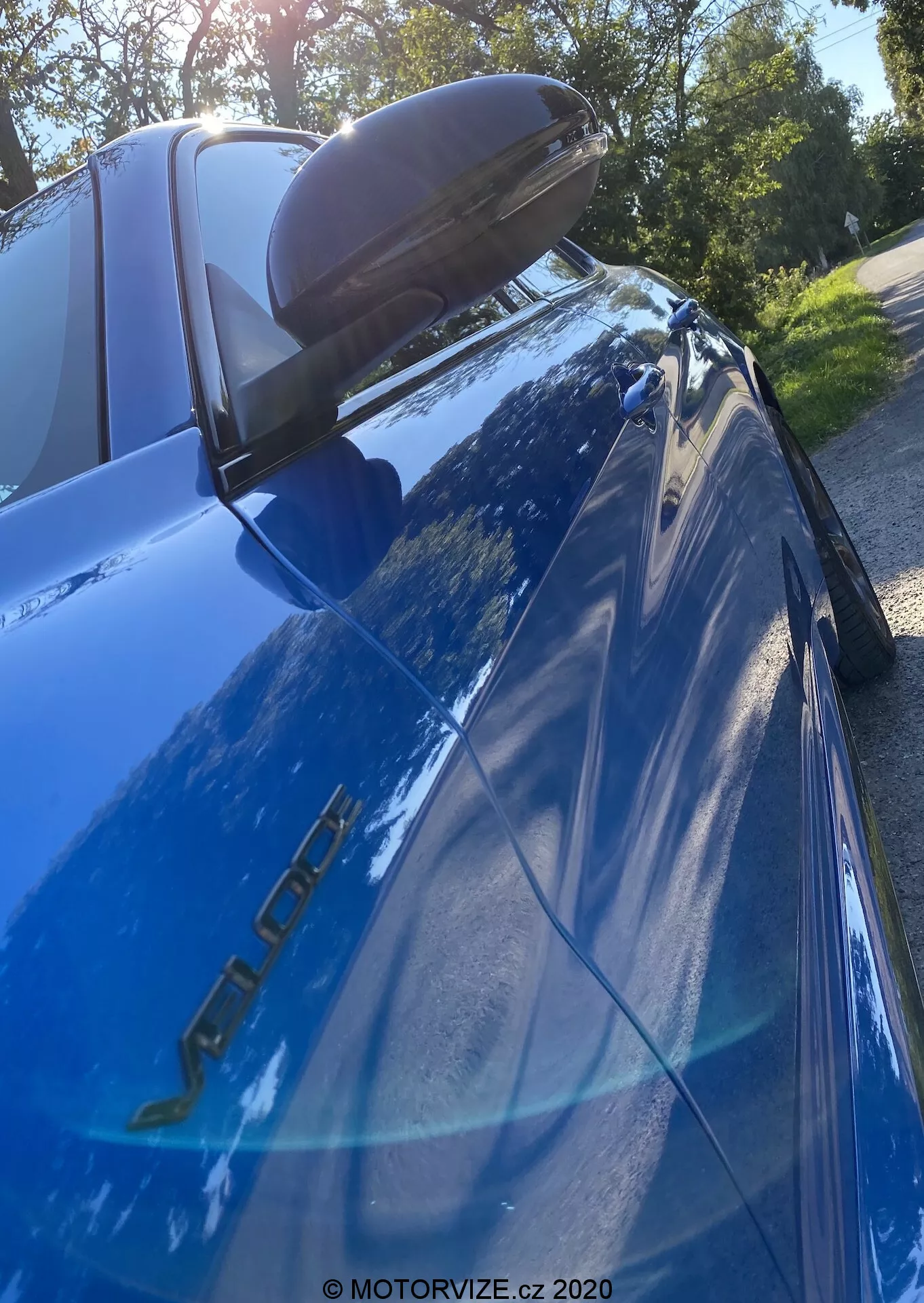 Imagen de primer plano de un Alfa Romeo Giulia azul (modelo 2016) fotografiado desde un ángulo bajo y hacia arriba centrado en la sección trasera. Se pueden ver las insignias del modelo en el lado del conductor, la pintura azul reflectante, la luz trasera LED izquierda, el alerón trasero negro, la ventana trasera inclinada con la tercera luz de freno, el reflejo de árboles y cielo brillante, y el espejo lateral con señal de giro integrada.