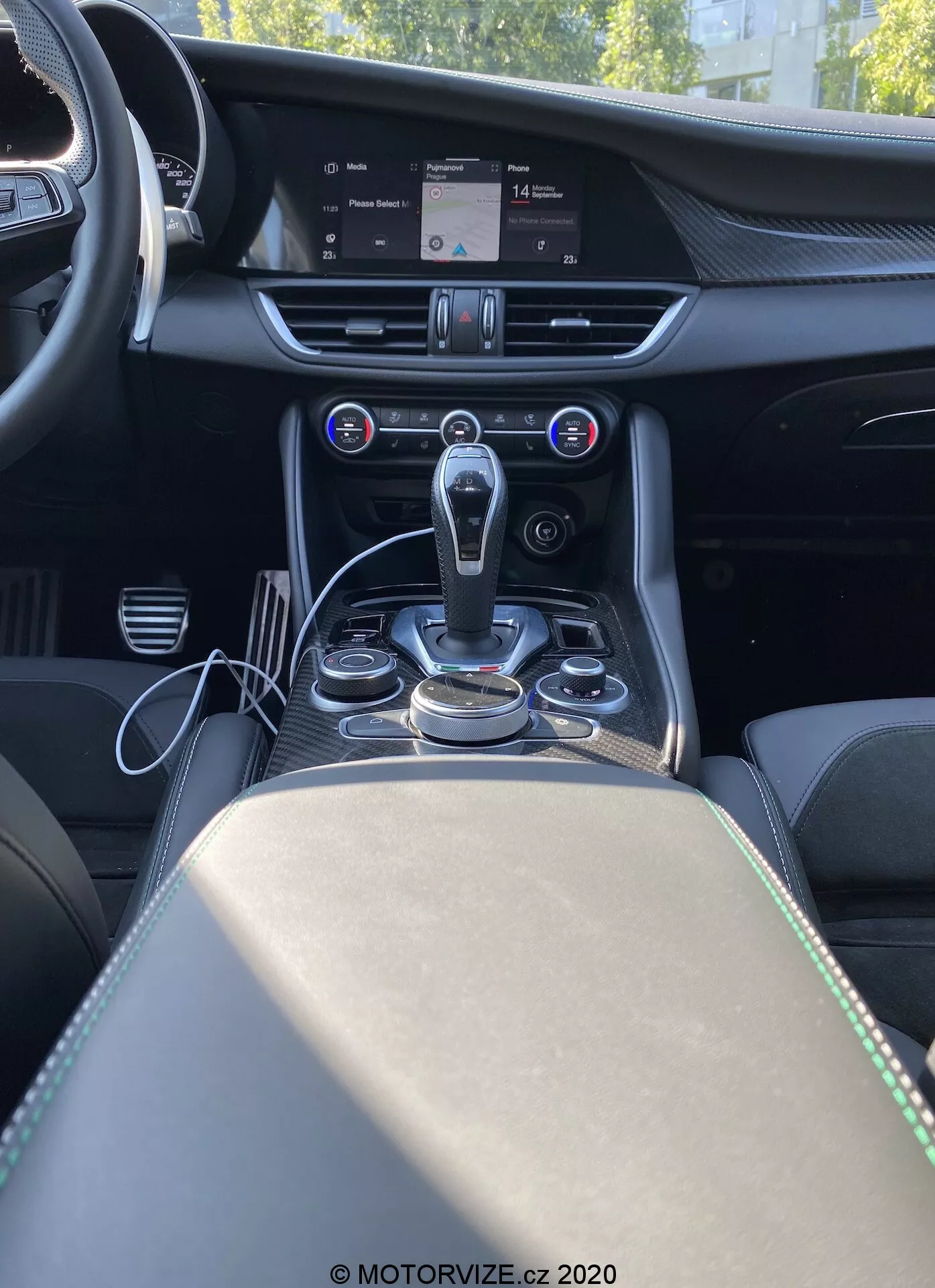 Imagen del interior del Alfa Romeo Giulia 2016 vista desde detrás de la consola central, mostrando la pila central, el túnel de la transmisión y parte del asiento del pasajero. La imagen ofrece una perspectiva frontal desde el punto de vista de un pasajero trasero, destacando una moderna palanca de cambios automática rodeada de diales metálicos y botones para el sistema de infoentretenimiento y modos de conducción. Encima del área de la palanca de cambios se encuentra el sistema de control climático automático de doble zona con una pantalla digital que muestra la configuración de la temperatura y botones para controlar la distribución de aire y la velocidad del ventilador. Una gran pantalla de infoentretenimiento en la parte superior de la pila central muestra el mapa del sistema de navegación. El tablero presenta detalles de costura y un acabado metálico que sugiere materiales premium. El reposabrazos y el compartimento de almacenamiento con dos portavasos son visibles detrás de la palanca de cambios, lo que mejora el diseño lujoso y enfocado en la tecnología de la cabina.