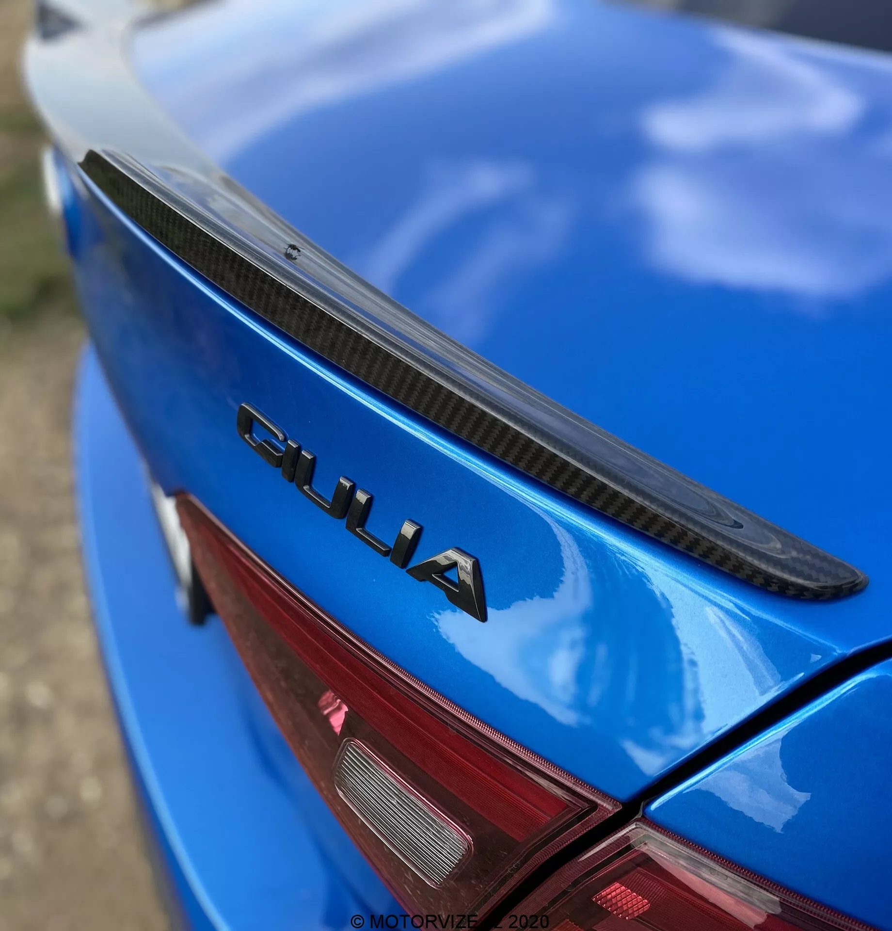 Vista de cerca y en ángulo de la sección trasera de un Alfa Romeo Giulia 2016 azul. La fotografía presenta la tapa del maletero con un alerón negro, mostrando pintura azul brillante y una prominente insignia cromada 'GIULIA'. También se ve una parte de la luz trasera LED roja. La insignia 'GIULIA' está enfocada con nitidez mientras que el alerón, la superficie del coche y la luz trasera se desdibujan gradualmente, destacando los elementos de marca y diseño del coche.