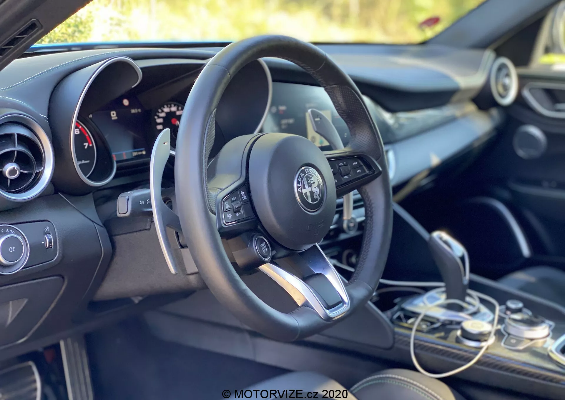 Vista interior de un Alfa Romeo Giulia 2016 centrada en el área del conductor y el tablero desde una perspectiva inclinada hacia la parte trasera y lateral izquierda. La configuración de conducción a la izquierda destaca un volante multifuncional moderno con controles incorporados, incluyendo botones para ajustar el volumen, acceder a menús y, probablemente, el control de crucero. El logo de Alfa Romeo es claramente visible en el centro del volante. Detrás del volante se observa parcialmente un panel de instrumentos mixto de displays analógicos y digitales, indicando un sistema de información configurable para el conductor. A la derecha del volante, apenas se ve la parte superior de la pantalla del sistema de infoentretenimiento, flanqueada por controles del sistema de clima y conmutadores funcionales debajo. El selector de cambios se encuentra en la parte inferior derecha, mostrando un diseño automático elegante. Los acabados de los materiales combinan tonos oscuros con acentos metálicos, reflejando el diseño interior premium del vehículo. La cabina está iluminada por luz natural, con un fondo verde que sugiere un entorno exterior o vegetativo.