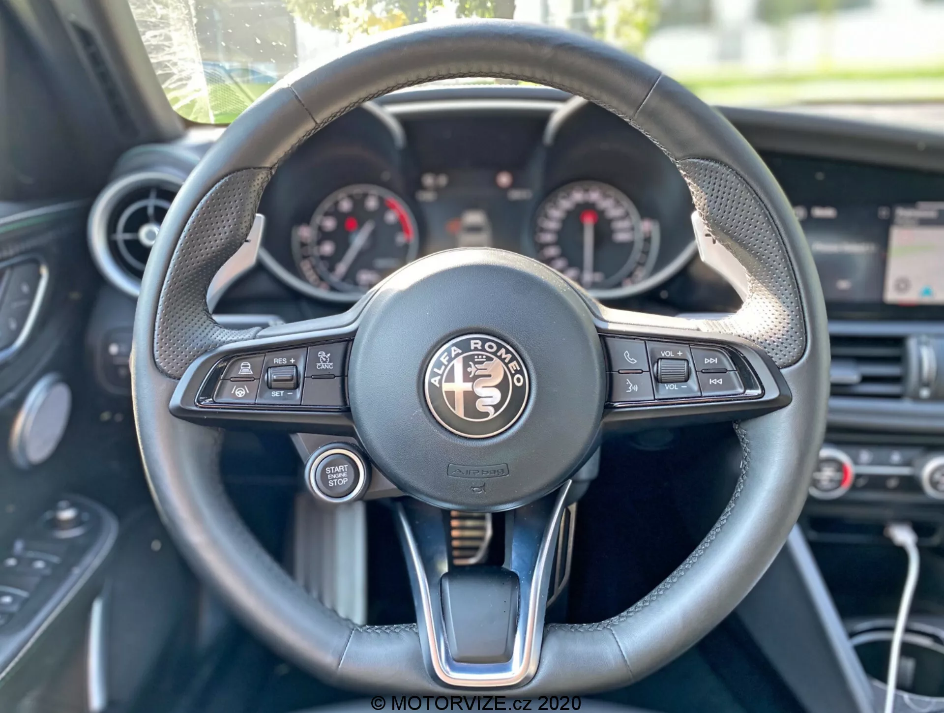 Perspectiva del conductor del interior del Alfa-Romeo Giulia 2016 centrada en el volante y el tablero. El volante multifuncional, ubicado en el centro, cuenta con botones para medios, control por voz, conectividad telefónica, información del vehículo y control de crucero. El panel de instrumentos presenta esferas analógicas, probablemente velocímetro y tacómetro, con una pantalla digital en el medio. El volante combina cuero, costuras y acentos metálicos, exudando lujo. En el fondo se ven los controles de clima y una pantalla central sobre ellos.