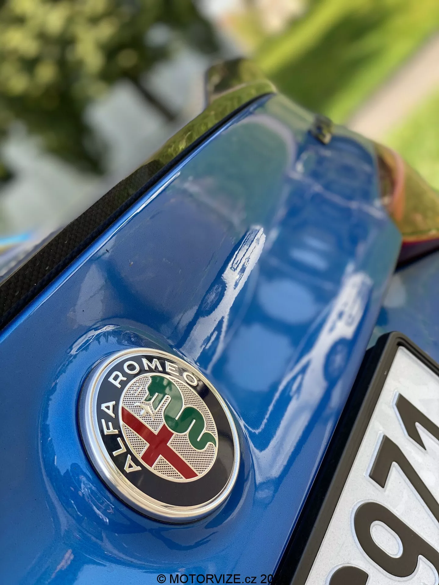 Vista cercana de la sección trasera del Alfa Romeo Giulia 2016 con el enfoque en el emblema de la marca desde un ángulo bajo. El distintivo, con una cruz roja sobre fondo blanco y una serpiente verde sobre fondo azul, está nítidamente enfocado en la tapa del maletero azul metálico, con detalles y reflejos de la superficie circundante.