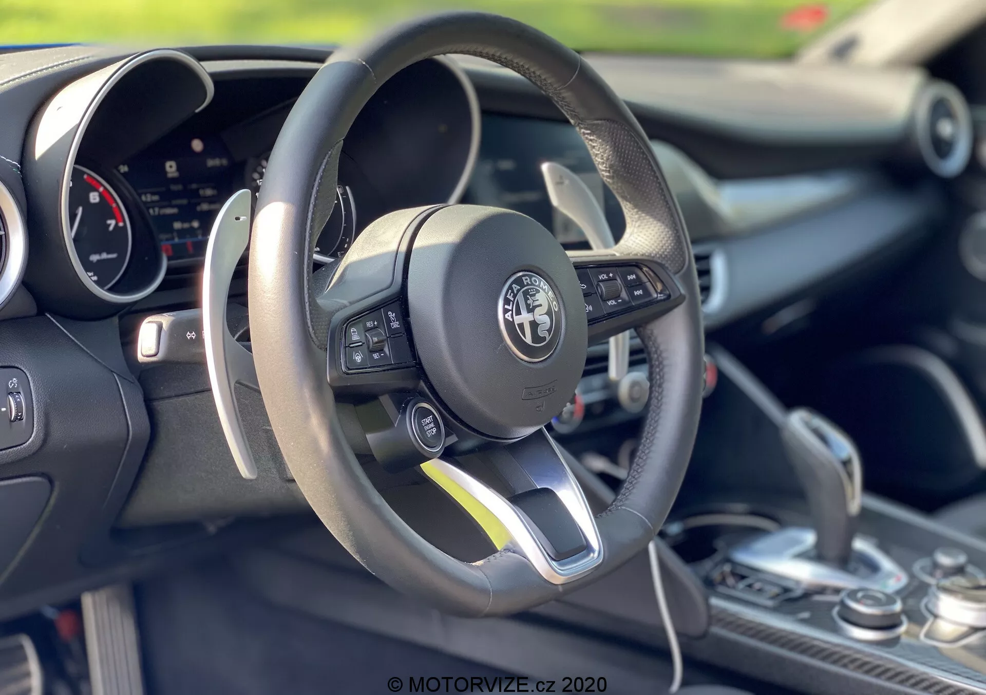 Toma interior de un Alfa-Romeo Giulia 2016 enfocándose en el área del conductor y el volante. El volante de fondo plano envuelto en cuero presenta controles multifunción en el lado izquierdo. Detrás del volante es visible un panel de instrumentos digital. La consola central muestra una palanca de cambios con una funda de cuero, lo que sugiere posibles capacidades de cambio manual. La imagen enfatiza el diseño deportivo del coche, la tecnología avanzada y la artesanía detallada con una profundidad de campo superficial que resalta los elementos del primer plano.