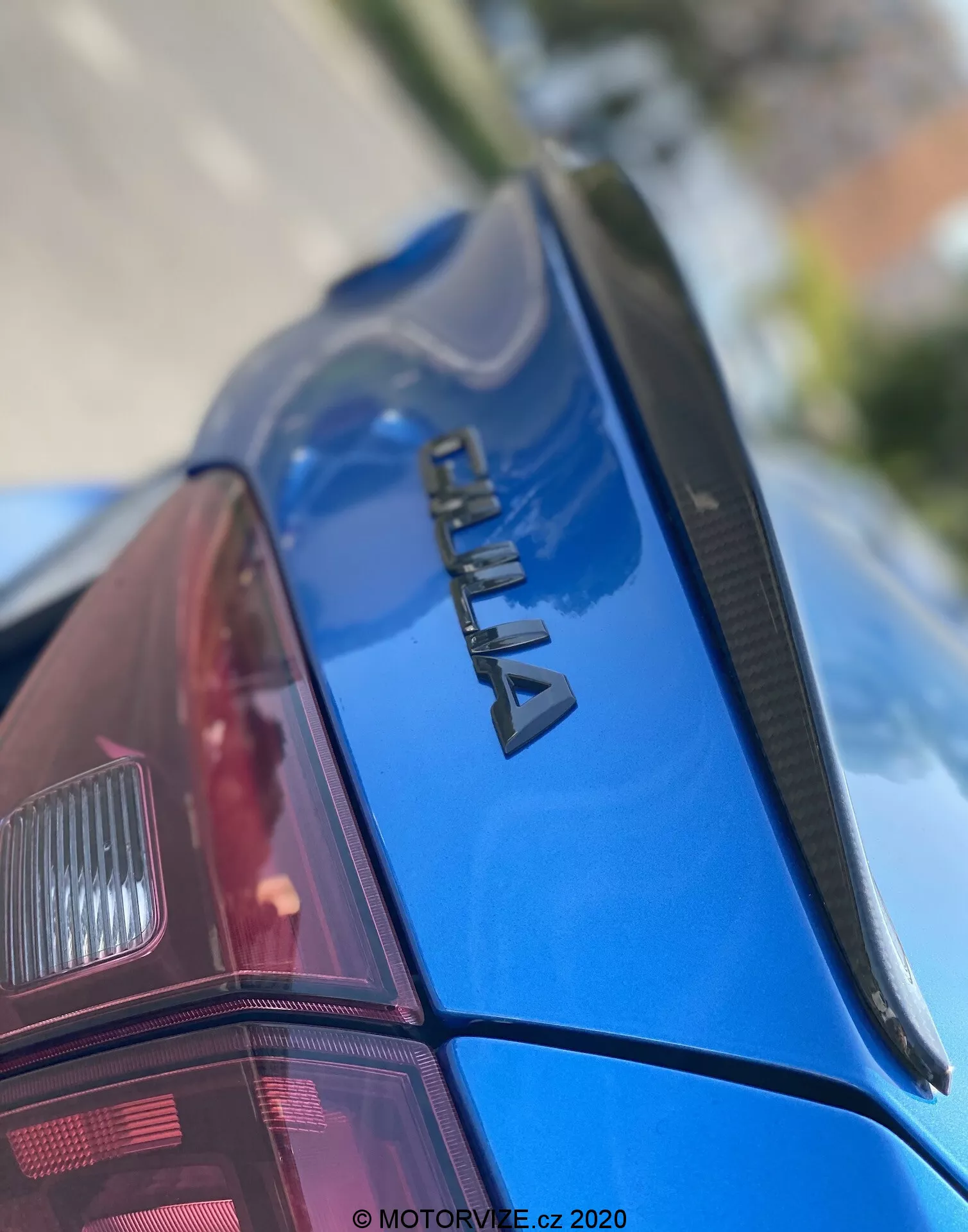 Imagen en primer plano de la sección trasera de un coche azul ŠKODA, vista desde un ángulo hacia arriba. El enfoque está en el logotipo de cromo 'ŠKODA' en la tapa del maletero, un spoiler estrecho y la pintura brillante y bien mantenida. El parabrisas trasero tiene líneas de desempañador y se ve parcialmente el conjunto de luces traseras modernas. La poca profundidad de campo enfatiza la marca, con la parte delantera y el fondo desenfocados. Con la marca de agua 'MOTORVIZE.cz 2020', indicando su fecha de publicación o captura.