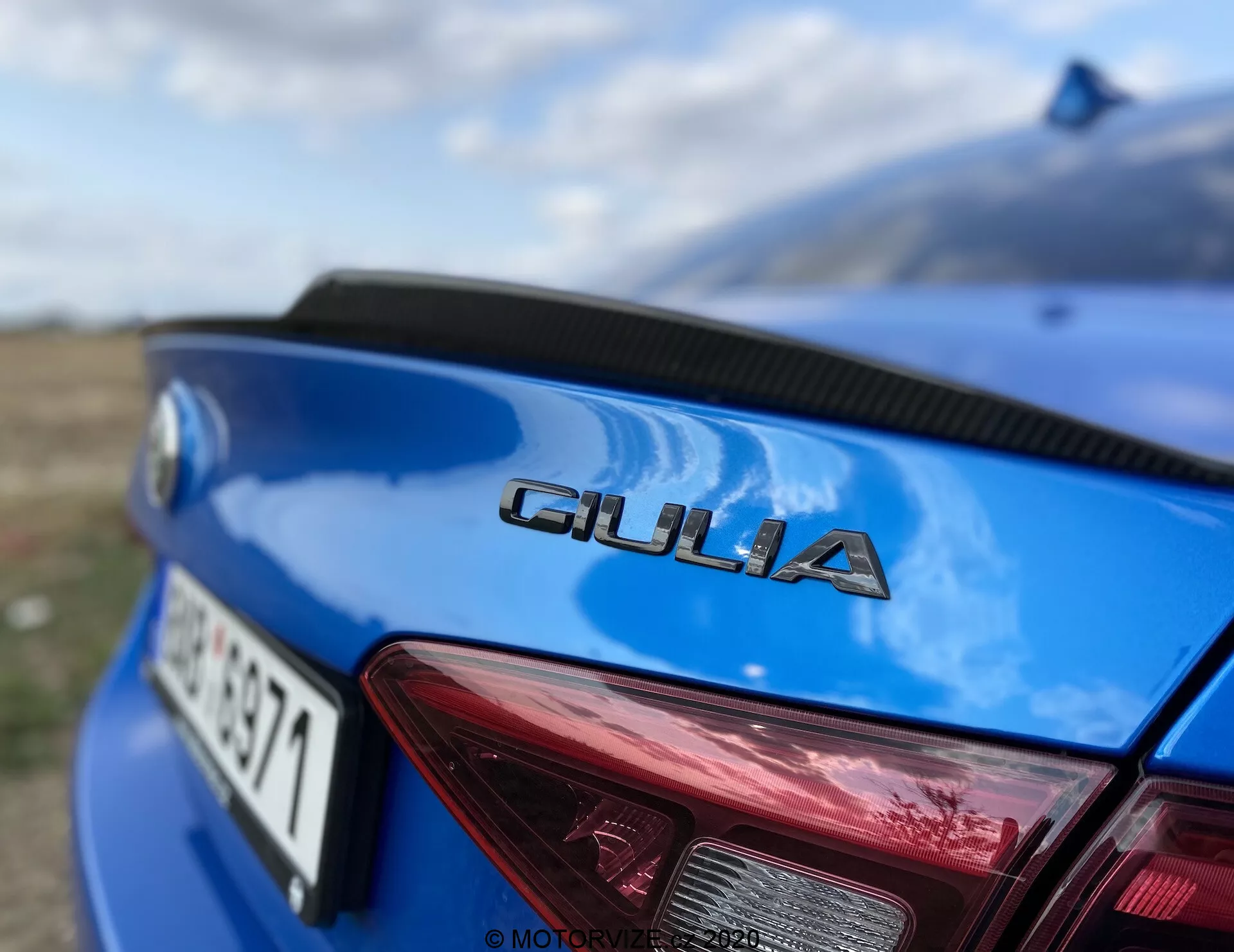 Primer plano de la parte trasera de un Alfa Romeo Giulia 2016 azul, tomada desde un ángulo bajo centrando en el distintivo cromado 'GIULIA' mostrado prominentemente en la tapa del maletero. Las letras nítidas contrastan con la pintura azul, destacando la tipografía de la marca. En la parte superior derecha se ve una porción segmentada de la luz trasera derecha, mostrando un diseño rojo distintivo con acentos blancos para las luces de reversa e intermitentes. La matrícula es parcialmente visible y está borrosa debajo de la designación del modelo. La curvatura del maletero sugiere un diseño aerodinámico, con un paisaje natural borroso en el fondo y un efecto bokeh de una apertura alta, enfatizando los detalles del vehículo.