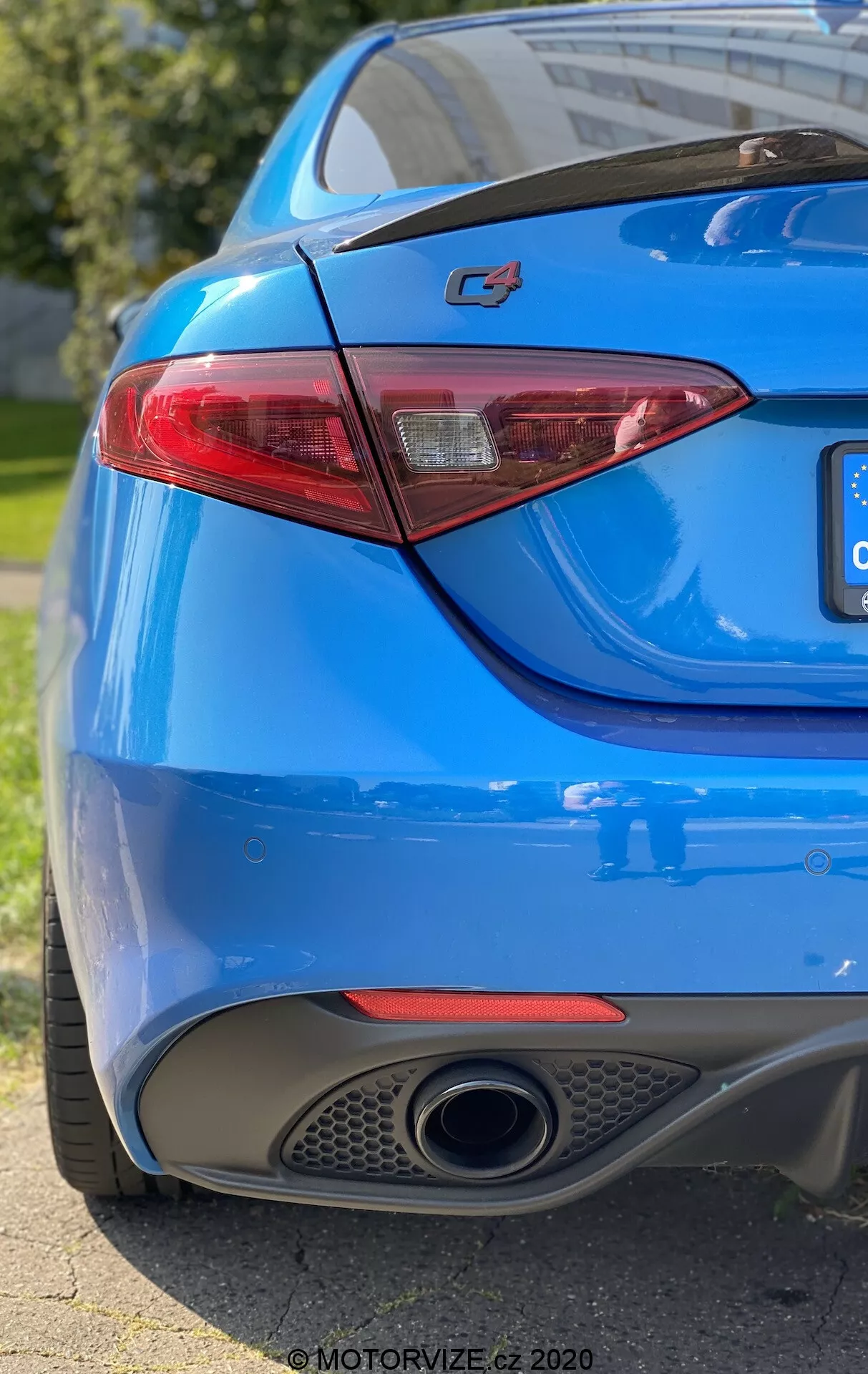 Un primer plano de la sección trasera de un coche azul que muestra el lado derecho desde un ángulo bajo tres cuartos. El encuadre captura el emblema 'C4' prominente en el maletero, una luz trasera LED elegante, pintura azul vibrante, una sola salida de escape cromada y el difusor integrado en el parachoques trasero. La poca profundidad de campo pone énfasis en el emblema y la luz trasera, mientras la luz natural del sol se refleja en las curvas suaves y deportivas de la carrocería del coche.