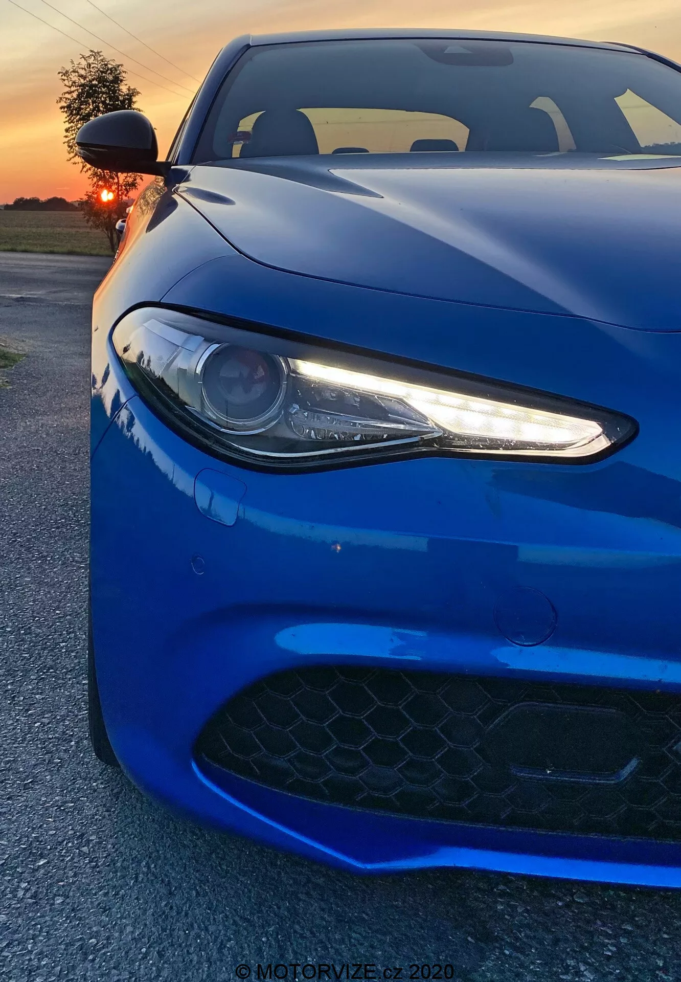 La imagen muestra un coche deportivo Alfa Romeo Giulia 2016 capturado desde un ángulo bajo frontal. El vibrante vehículo azul profundo llena el encuadre, enfatizando su faro izquierdo con luces diurnas LED, una parte del capó y la rejilla de aire en el parachoques delantero. El faro de tipo proyector, con sus elegantes acentos LED blancos, proporciona un aspecto agresivo y moderno. La rejilla negra de patrón hexagonal sugiere capacidades de alto rendimiento. La toma captura las líneas y contornos aerodinámicos del coche, con reflejos del atardecer que realzan su acabado brillante. En el fondo se ve un cielo iluminado en naranja parcialmente oculto por el sol. El ángulo cercano del fotógrafo muestra detalles de diseño, evocando una sensación de poder y elegancia.