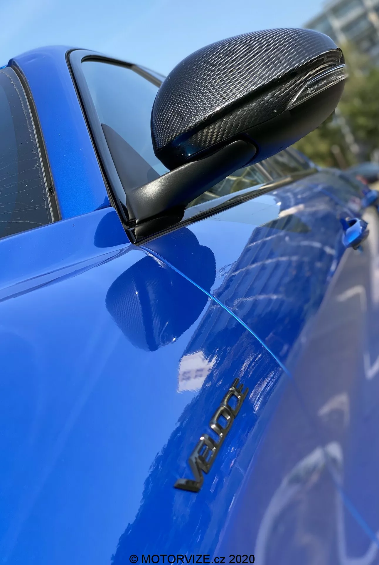 Imagen de primer plano de un automóvil Alfa Romeo Giulia 2016 azul, enfatizando el espejo retrovisor lateral con un acabado de fibra de carbono en una carcasa aerodinámica, montado en un soporte negro. La foto se enfoca nítidamente en el espejo, la puerta del conductor y la insignia metálica 'VERON', destacando estos detalles con poca profundidad de campo y una superficie brillante y bien mantenida.