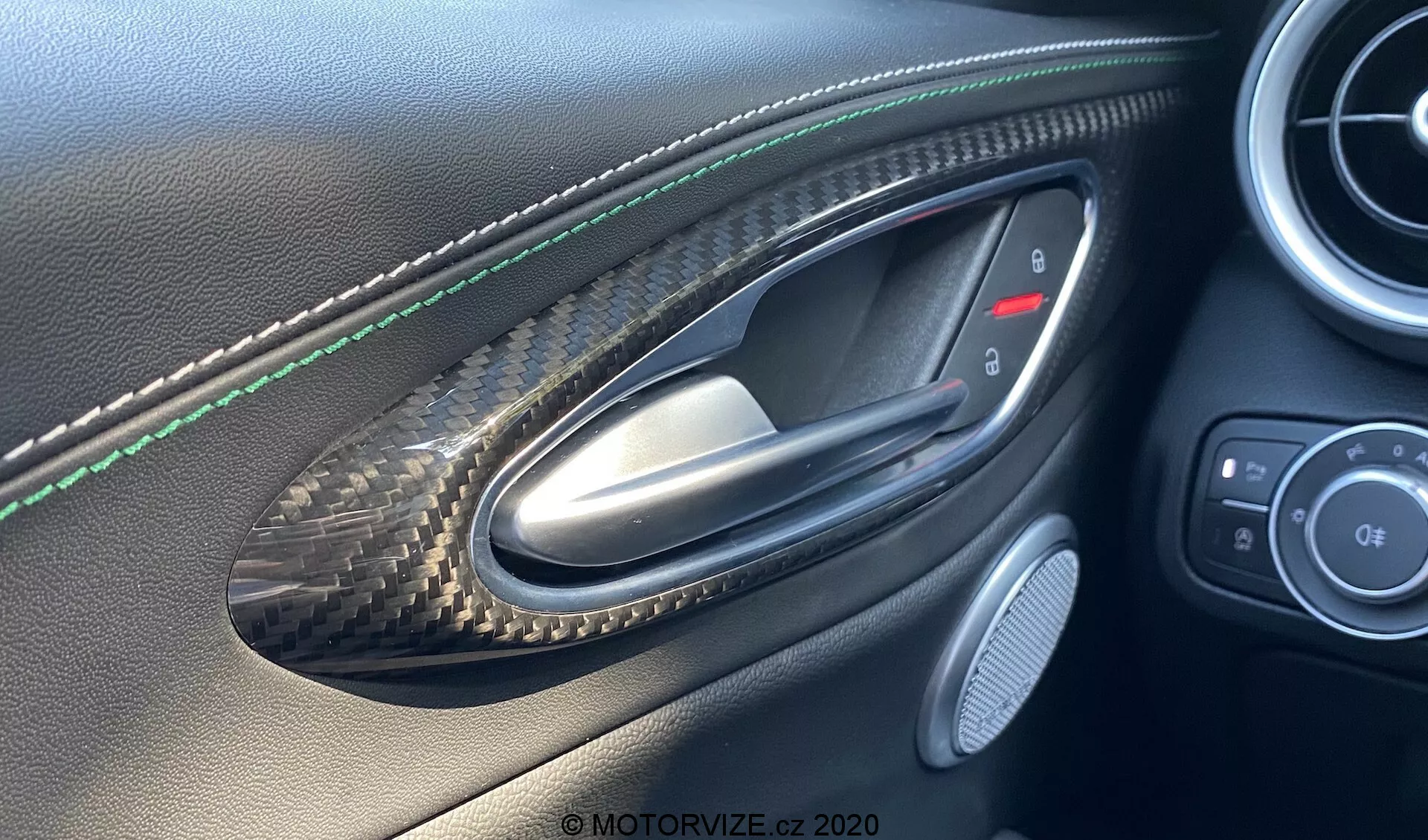 Imagen interior del lado del conductor del Alfa Romeo Giulia 2016 vista desde la perspectiva de alguien sentado en el interior, destacando un tirador de puerta metálico colocado en una hendidura con patrón de fibra de carbono, costuras verdes en el panel de cuero de la puerta, botones de control para espejos laterales y ventanas, una rejilla de altavoz y acabados de cuero de alta calidad y fibra de carbono indicativos de un vehículo de lujo o rendimiento.