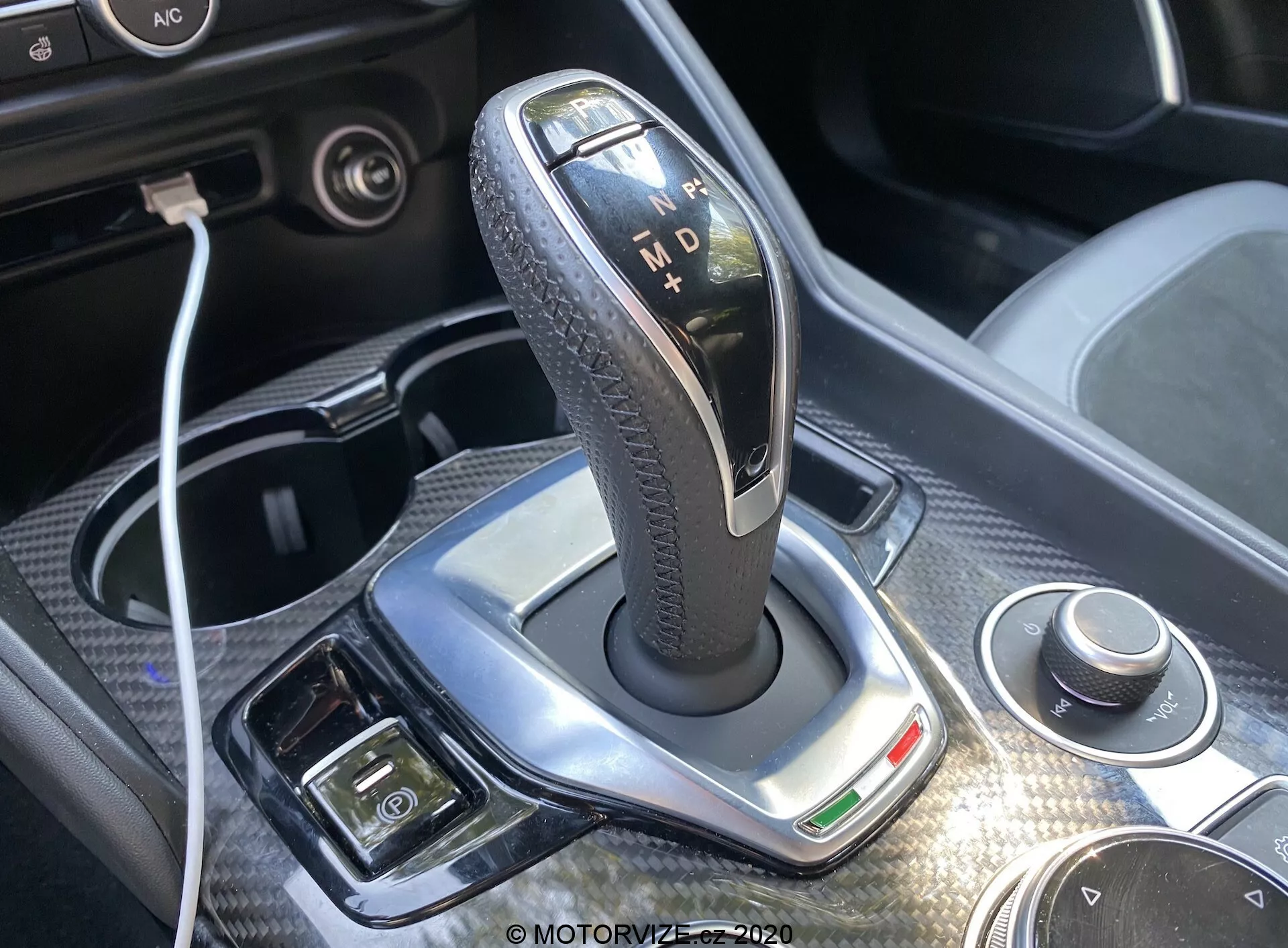 Primer plano de la palanca de cambios de la transmisión automática del Alfa-Romeo Giulia 2016, con un elegante borde plateado pulido, empuñadura negra texturizada de cuero e indicadores iluminados para 'P', 'R', 'N', 'D' y 'M'. La luz diurna brillante resalta el diseño, con un dial giratorio y conectividad USB visibles cerca. Acabado cromado, luz indicadora verde y botones adicionales para freno de estacionamiento electrónico y control de clima son visibles, ofreciendo una visión del lujo y tecnología avanzada del interior del coche.