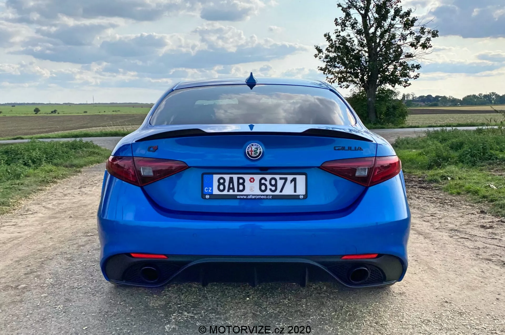Vista trasera de un sedán Alfa Romeo Giulia 2016 azul brillante, tomada desde una perspectiva a nivel de los ojos. La imagen muestra todo el trasero del coche, centrado en el marco, con la matrícula, las luces traseras y el logotipo del fabricante claramente visibles. Las características incluyen un sistema de escape doble, luces traseras modernas y elegantes, un elemento de difusor en el parachoques, un alerón en la tapa del maletero, sensores de estacionamiento y un limpiaparabrisas trasero, lo que sugiere un equilibrio de carácter deportivo, seguridad y practicidad. El escenario es una zona rural con un paisaje plano y un cielo parcialmente nublado durante el día.