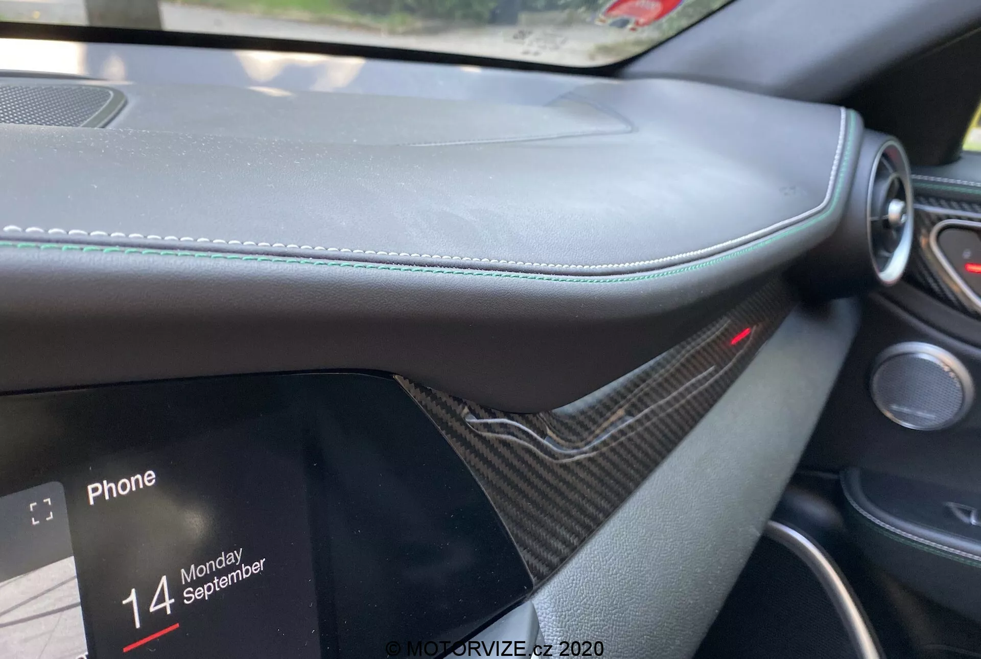 Detalle interior del Alfa Romeo Giulia 2016 desde la perspectiva del pasajero, centrado en el salpicadero con material similar al cuero y costuras visibles, moldura similar a la fibra de carbono, pantalla de infoentretenimiento parcialmente visible mostrando opciones de conectividad telefónica con el texto 'Phone' y 'Monday 14 September'. Luz roja encima del sistema de infoentretenimiento indicando un sistema de seguridad o protección activo, enfatizando el lujo y el diseño moderno.