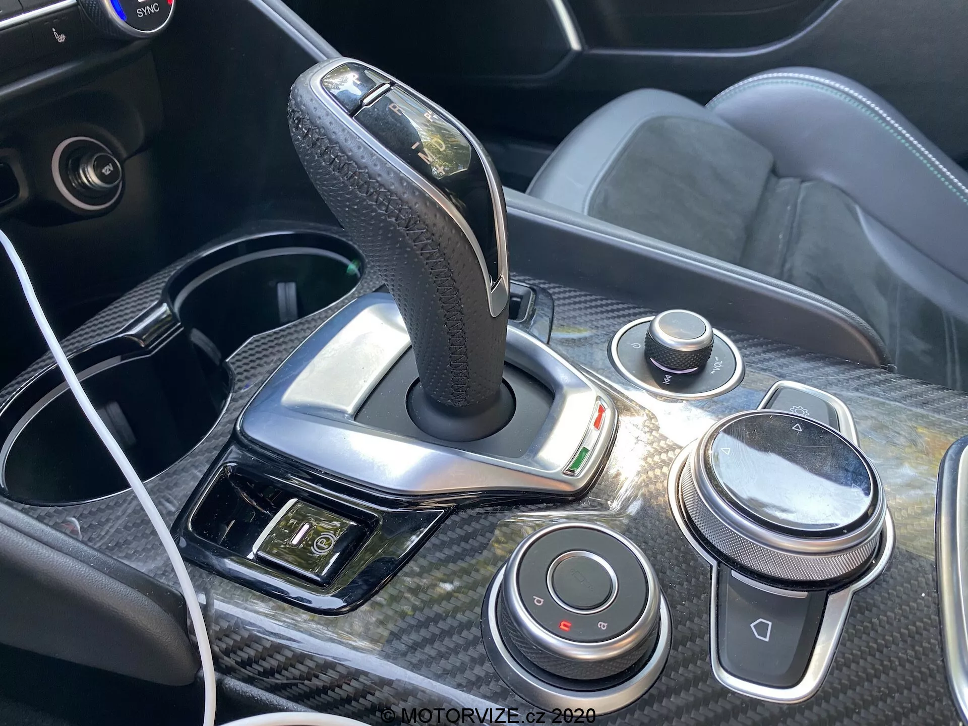 Vista de cerca de la consola central y área de la palanca de cambios del Alfa Romeo Giulia 2016, con una palanca de cambios automática envuelta en cuero con detalles cromados, rodeada de un acabado de fibra de carbono. La consola incluye un botón de control tipo iDrive, dial de control de volumen, botones para dinámica de conducción, puerto USB con cable conectado y un panel con el control de luces de emergencia y otros, enfatizando el diseño ergonómico y tecnológico del interior del coche.