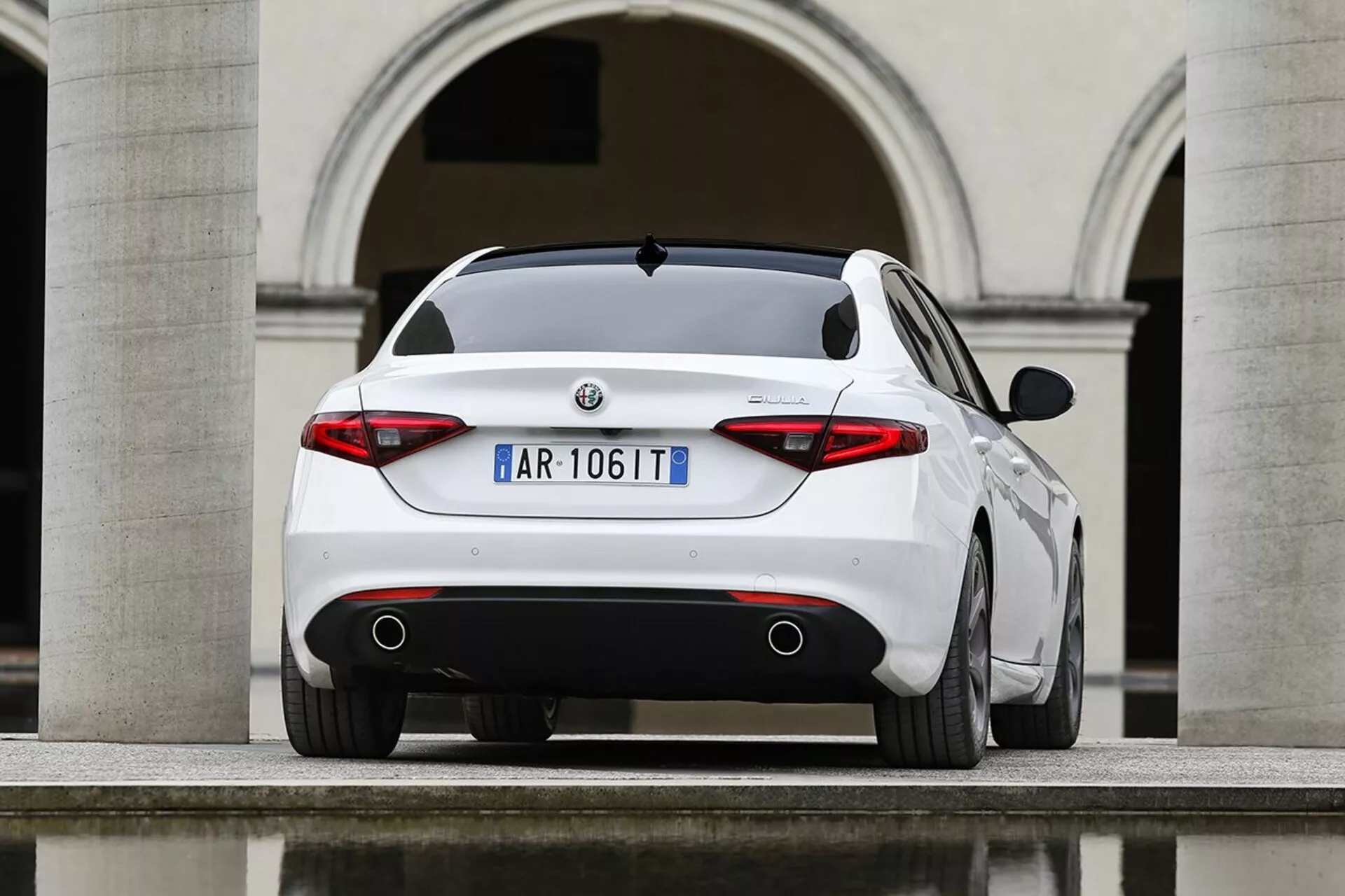 Alfa Romeo Giulia blanco del 2016 fotografiado desde un ángulo trasero de tres cuartos destacando su perfil trasero deportivo, luces traseras distintivas, tubos de escape dobles y características de diseño elegante contra un telón de fondo de arquitectura europea