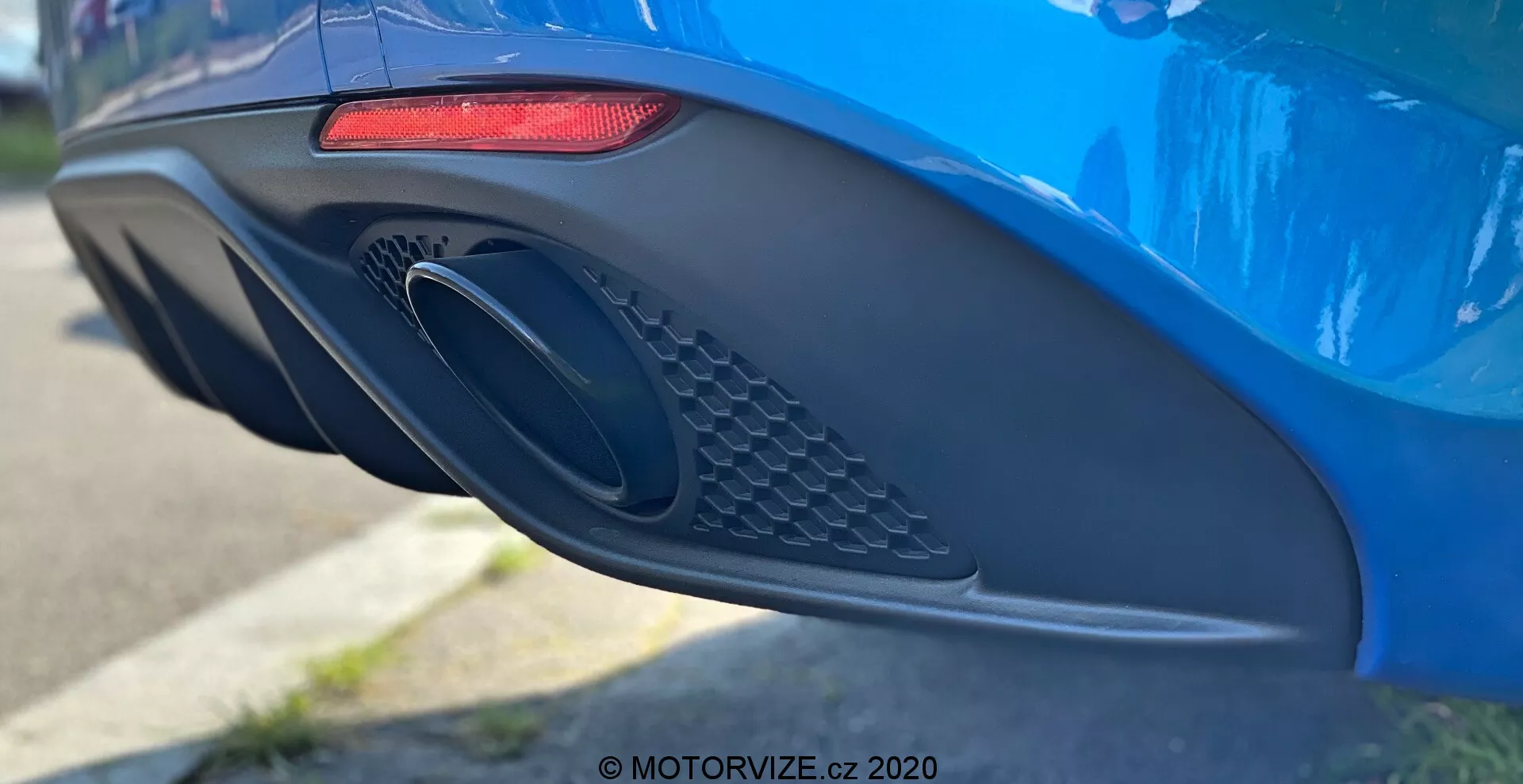 Vista lateral en primer plano del parachoques trasero y el área del escape de un vibrante Alfa Romeo Giulia 2016 azul, que muestra sus características de diseño moderno, incluyendo una luz trasera LED horizontal alargada roja, un gran puerto de escape ovalado con detalle de malla, y un parachoques aerodinámico con varias curvas e indentaciones, fotografiado con poca profundidad de campo para resaltar los detalles del coche.