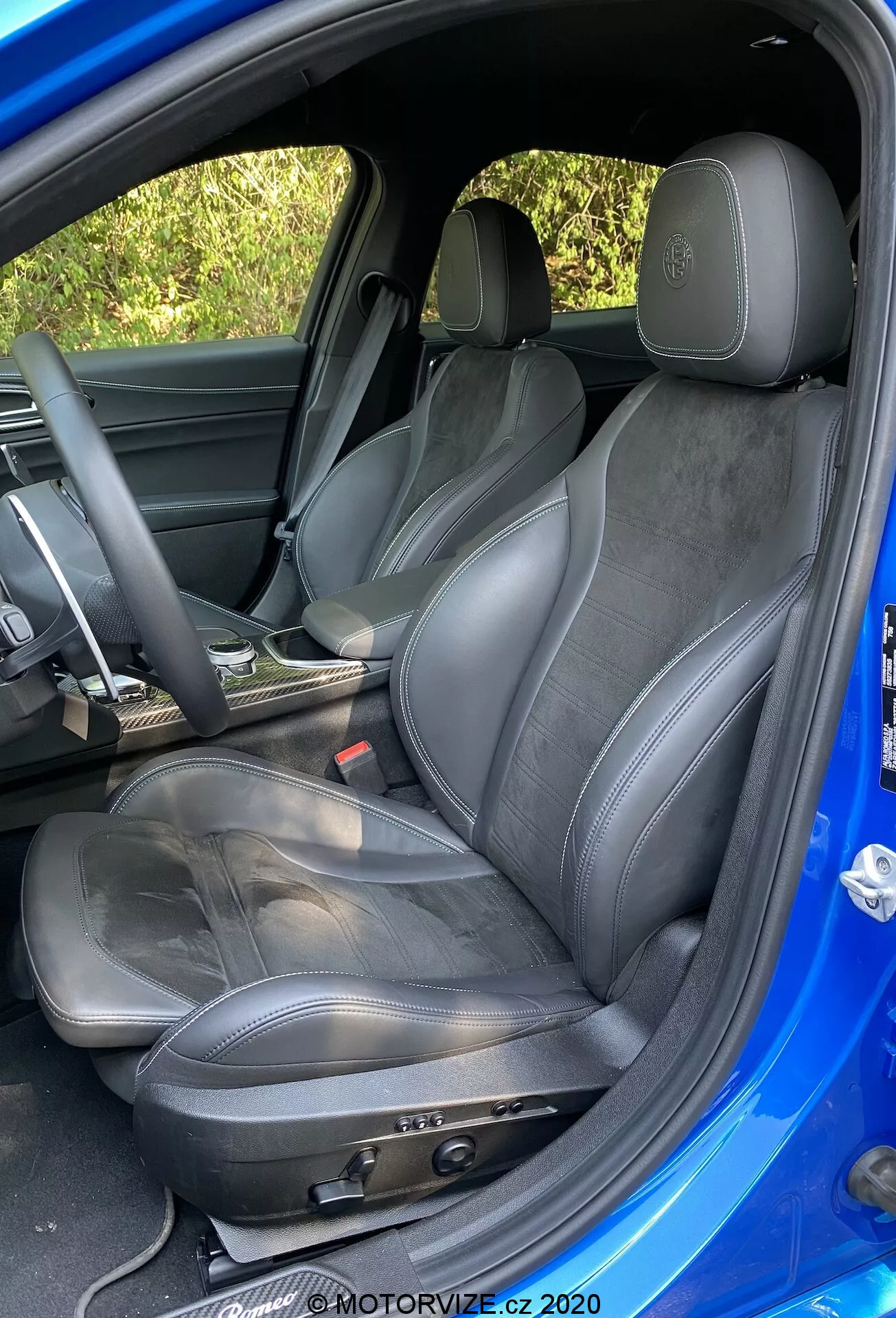 Vista detallada del asiento del conductor deportivo en un Alfa-Romeo Giulia 2016, mostrando ajustes manuales del asiento, tapicería de cuero y gamuza, logo en relieve en el reposacabezas, ligero desgaste, volante parcialmente visible envuelto en cuero y revestimiento interior de la puerta a juego, capturado desde un ángulo exterior por encima del nivel del asiento con interior oscuro y exterior azul brillante contrastante.