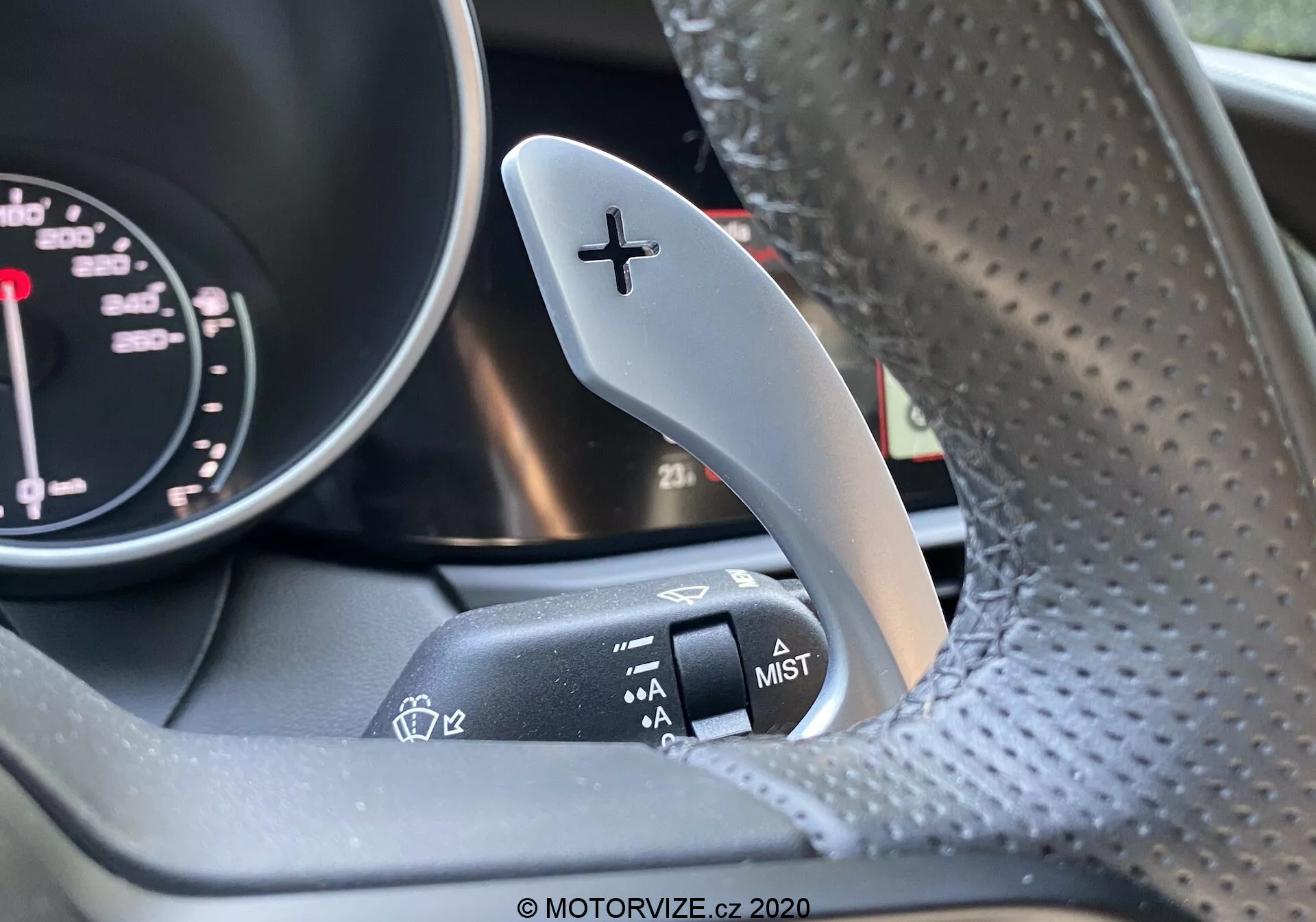 Toma interior de cerca de un Alfa Romeo Giulia 2016, centrada en la palanca de cambios del lado derecho con un signo '+' para subir de marcha, y la palanca de control del limpiaparabrisas adyacente en la columna de dirección. Parte del tacómetro del conjunto de instrumentos es ligeramente visible. El volante, hecho de cuero perforado, está algo desenfocado. La imagen resalta el contraste entre la palanca de cambios metálica y la palanca de control del limpiaparabrisas con acabado mate.