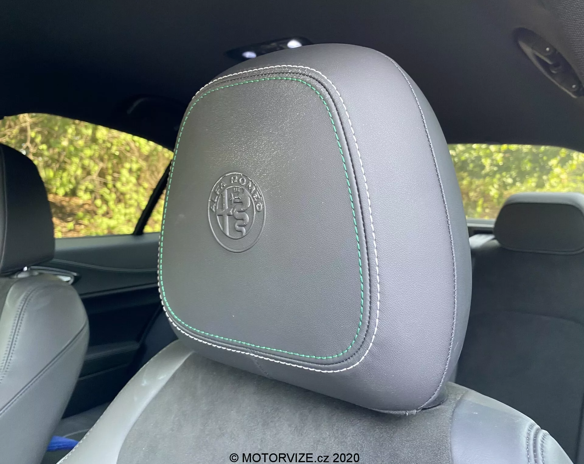 Primer plano de un reposacabezas del coche en el Alfa Romeo Giulia 2016, mostrando una tapicería de cuero gris claro con costuras en contraste de color verde claro, visto desde el asiento trasero. El reposacabezas tiene un logo circular en relieve que sugiere un acabado premium o personalizado. El fondo muestra follaje borroso a través de las ventanas del coche, insinuando un entorno al aire libre y con luz de día.