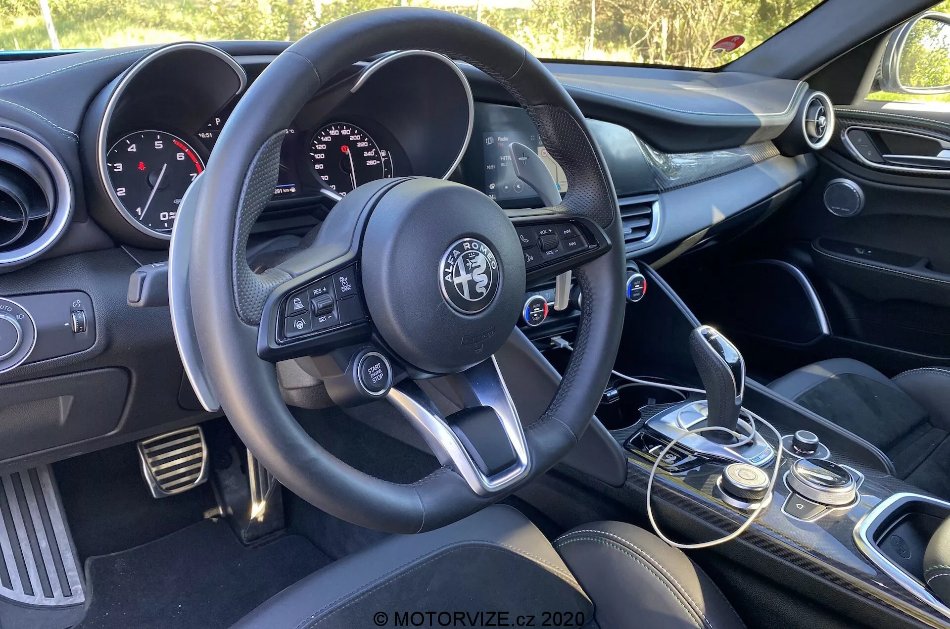 Vista interior del Alfa Romeo Giulia 2016 enfocada en el lado del conductor desde la perspectiva del pasajero, mostrando el volante de tres radios con botones multifuncionales, el logotipo del fabricante en el centro, el panel de instrumentos con diales analógicos y pantalla digital, la consola central con controles de audio y climatización, la palanca de cambios manual con funda de cuero, el selector de modos de conducción, los asientos delanteros con tapicería oscura y el panel de la puerta del conductor parcialmente visible con controles de ventanas y espejos.