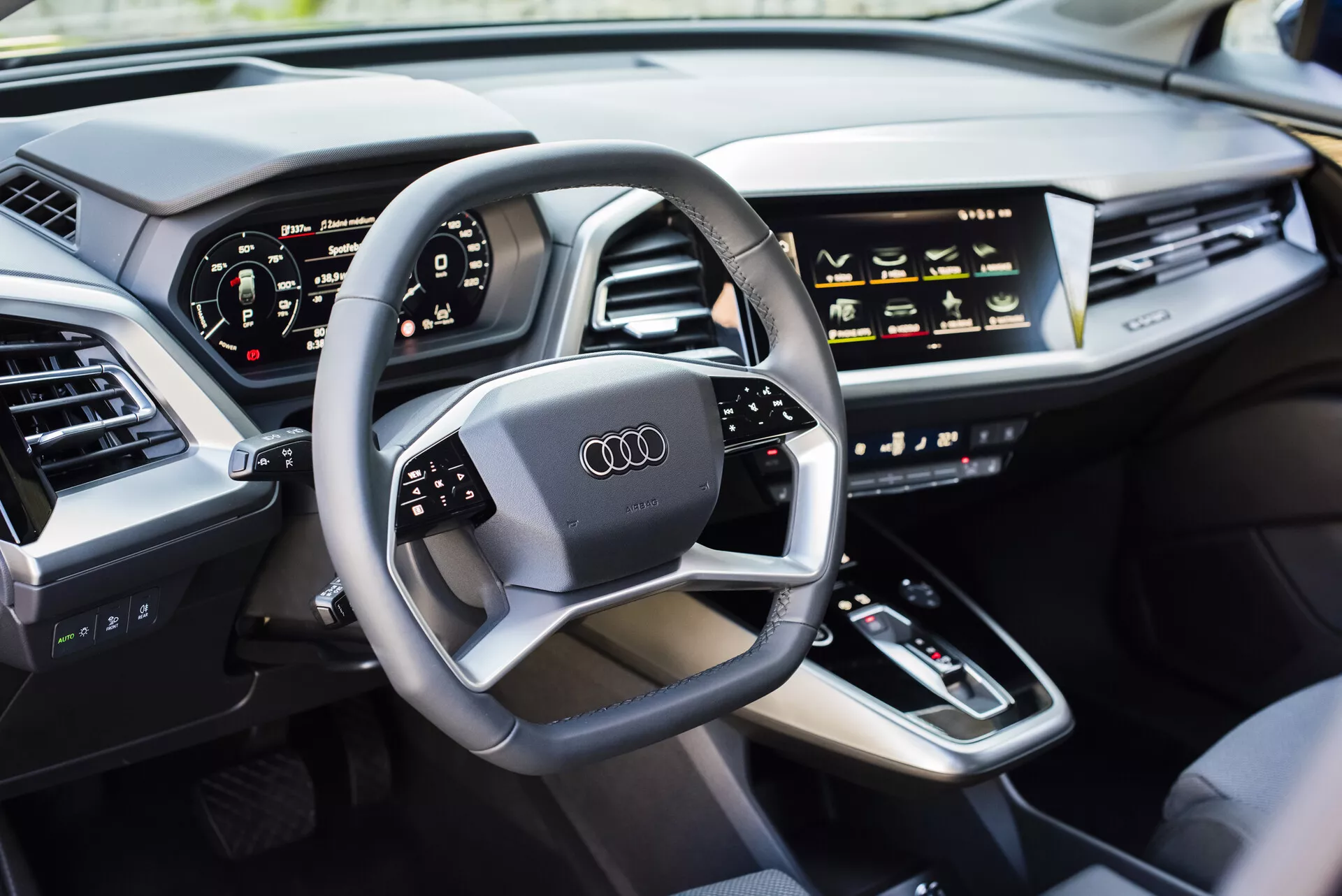 Vista interior de un Audi Q4 e-tron 2021 desde el lado del conductor, mostrando un volante multifuncional con el logo de Audi, panel de instrumentos digital, consola central con palanca de cambios, controles táctiles HVAC, pantalla de infotenimiento, tablero elegante con ventilaciones de aire integradas, y una puerta acolchada de cuero de lujo, exhibiendo la tecnología moderna de los coches y las características de los vehículos de lujo.