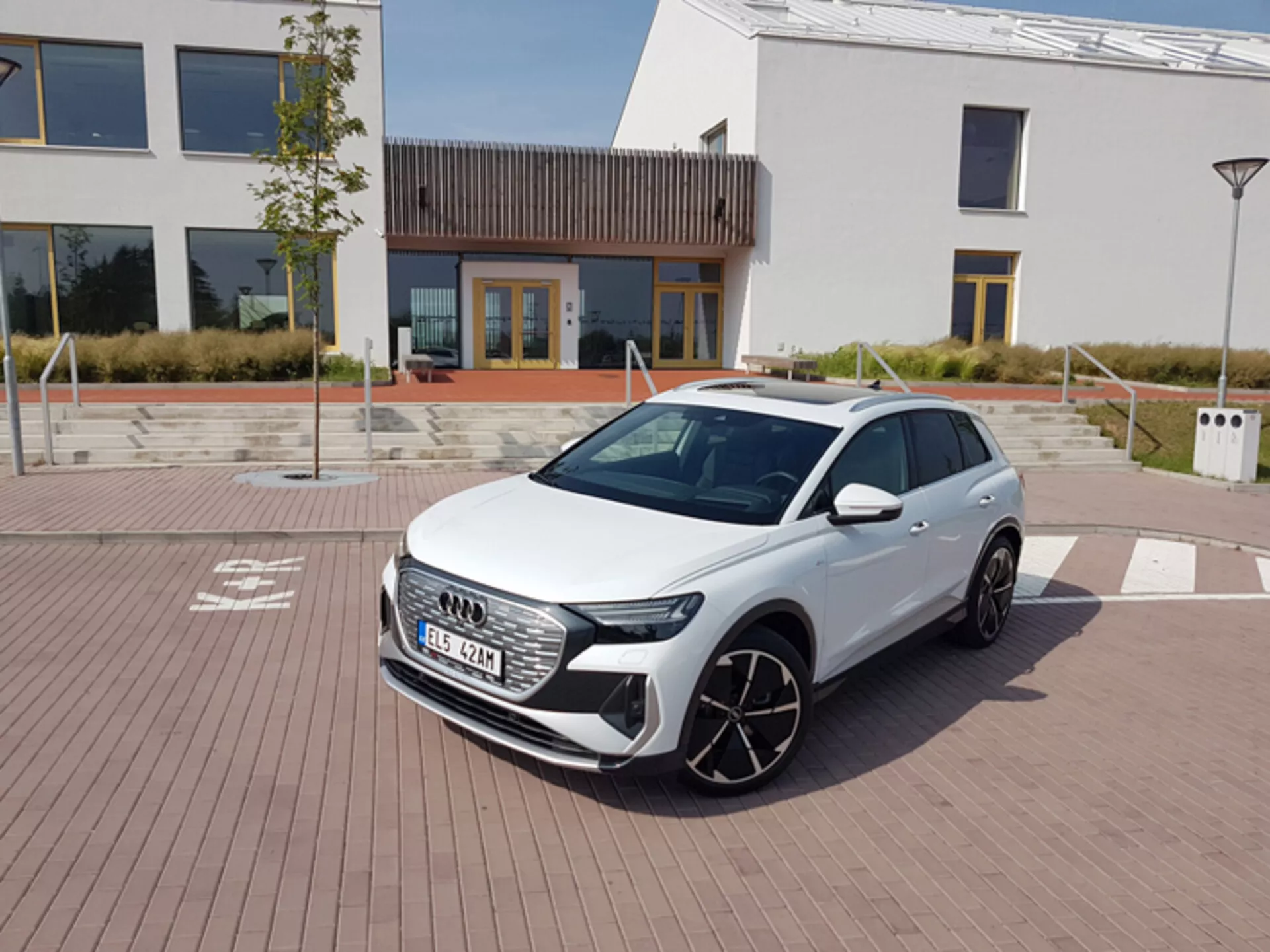 Audi Q4 e-tron 2021 en blanco, mostrando sus modernos faros LED, parrilla frontal hexagonal y grandes ruedas de aleación, aparcado en un pavimento con patrón de ladrillo con arquitectura urbana y una estación de carga en el fondo, bajo un cielo claro al mediodía.