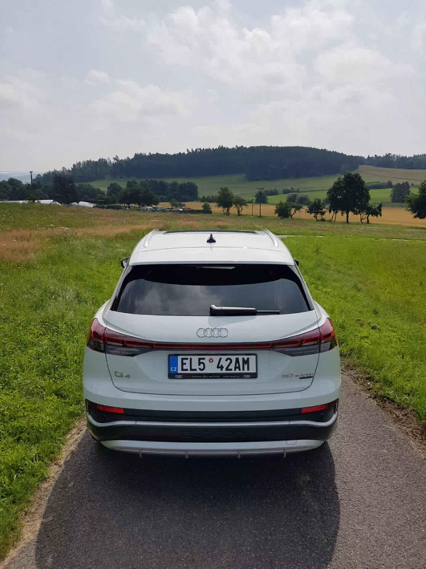 Vista trasera de un Audi Q4 e-tron 2021 plateado en un camino pavimentado estrecho en medio de un paisaje pastoril con campos y árboles bajo un cielo parcialmente nublado, destacando su elegante diseño, barra de luz de freno, spoiler trasero, sensores de estacionamiento traseros y ruedas de aleación de dos tonos.