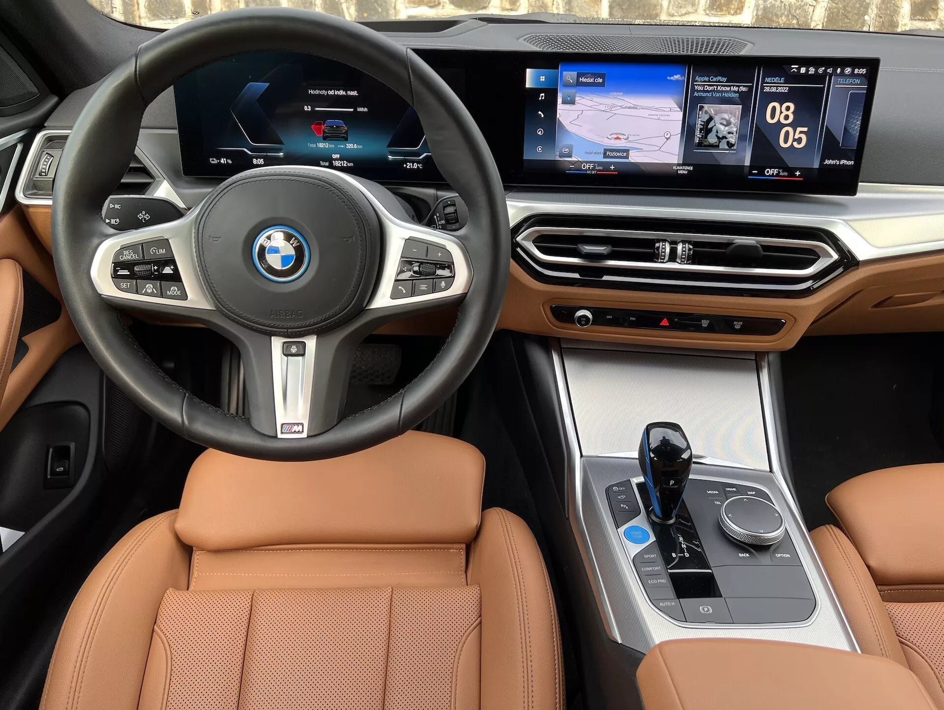Vista interior de un BMW i4 2021, con volante central con logo de BMW, botones multifuncionales, cuadro de instrumentos digital, sistema de infoentretenimiento central sensible al tacto que muestra navegación y medios, acentos de madera de lujo en el tablero, interfaz de control del clima, selector de marchas electrónico, controlador iDrive y asientos de cuero color canela.