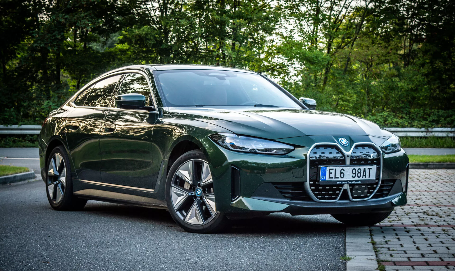 Sedán BMW i4 verde de cuatro puertas del 2021 aparcado en una calle de adoquines junto a una zona con césped y follaje al fondo, con una vista en ángulo frontal que muestra la gran parrilla del coche, faros LED, entradas de aire y llantas de aleación