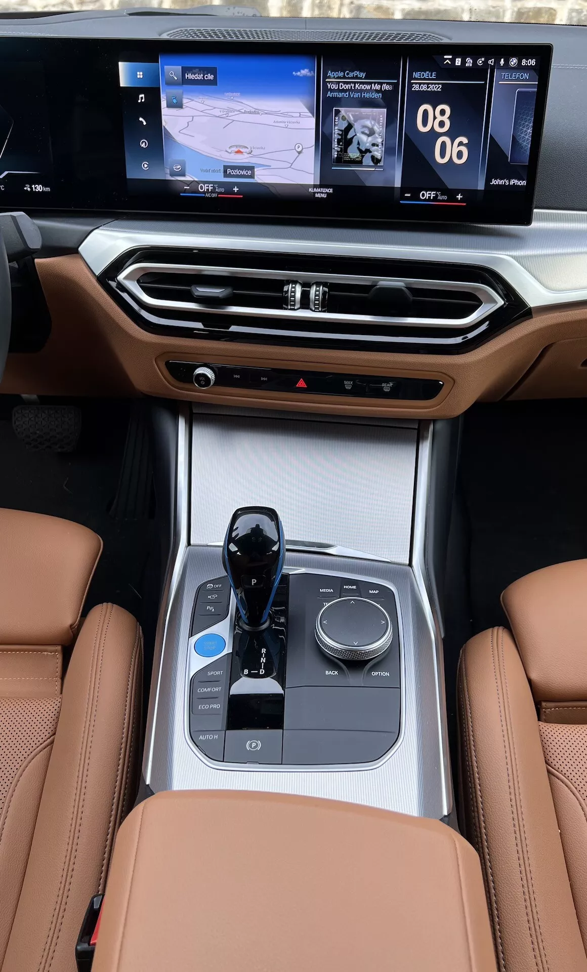 Vista del centro de mando interior de un BMW i4 2021 desde los asientos traseros, destacando los asientos de cuero, selector de marchas electrónico, perilla de control multimedia, portavasos, controles de clima y pantalla de infoentretenimiento de doble interfaz con navegación y pantallas de medios
