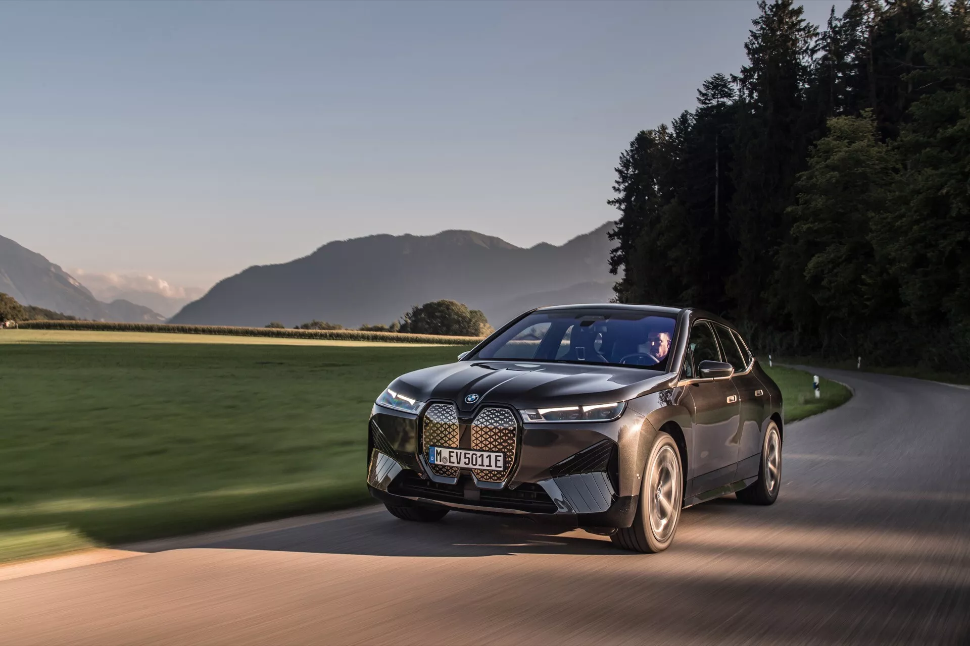 SUV eléctrico moderno BMW iX 2021 (i20) en movimiento en una carretera pavimentada al amanecer o al atardecer, mostrando su trabajo de pintura en dos tonos, diseño elegante con una parrilla prominente, faros distintivos y ruedas aerodinámicas contra un telón de fondo pintoresco.