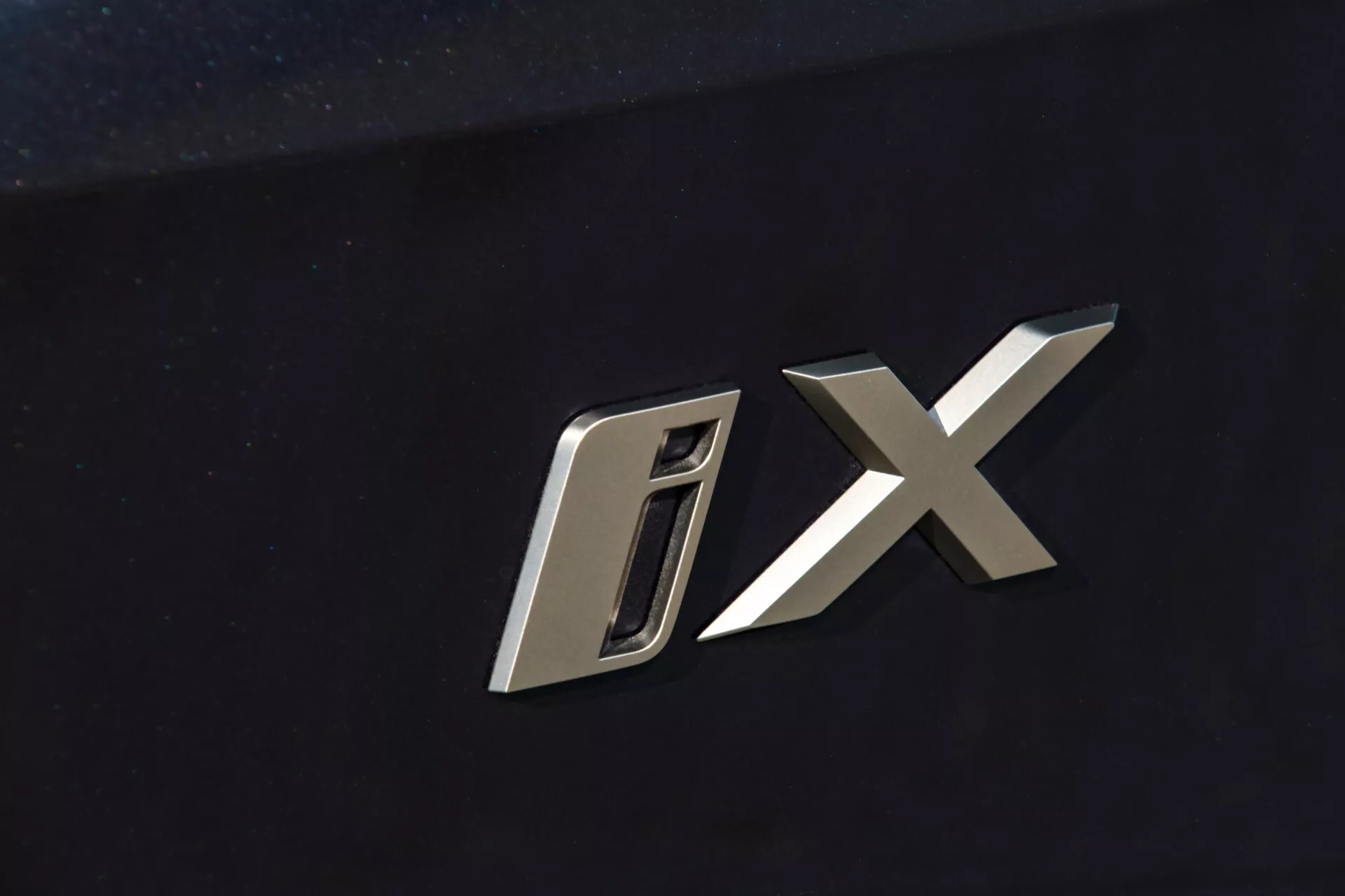 Vista de cerca del emblema del modelo BMW iX 2021 con caracteres 'D' y 'X' metalizados estilizados contra un fondo oscuro de carrocería