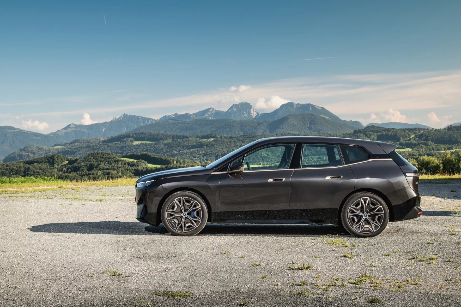 Perfil lateral de un moderno SUV BMW iX (i20) del 2021 sobre grava con fondo montañoso bajo un cielo azul claro, con grandes ruedas de aleación de múltiples radios, diseño elegante con línea de cintura alta y techo inclinado, vidrio de privacidad, señales de giro integradas en espejos del color del cuerpo, parrilla frontal visible y grupo de faros LED, molduras cromadas y techo panorámico.