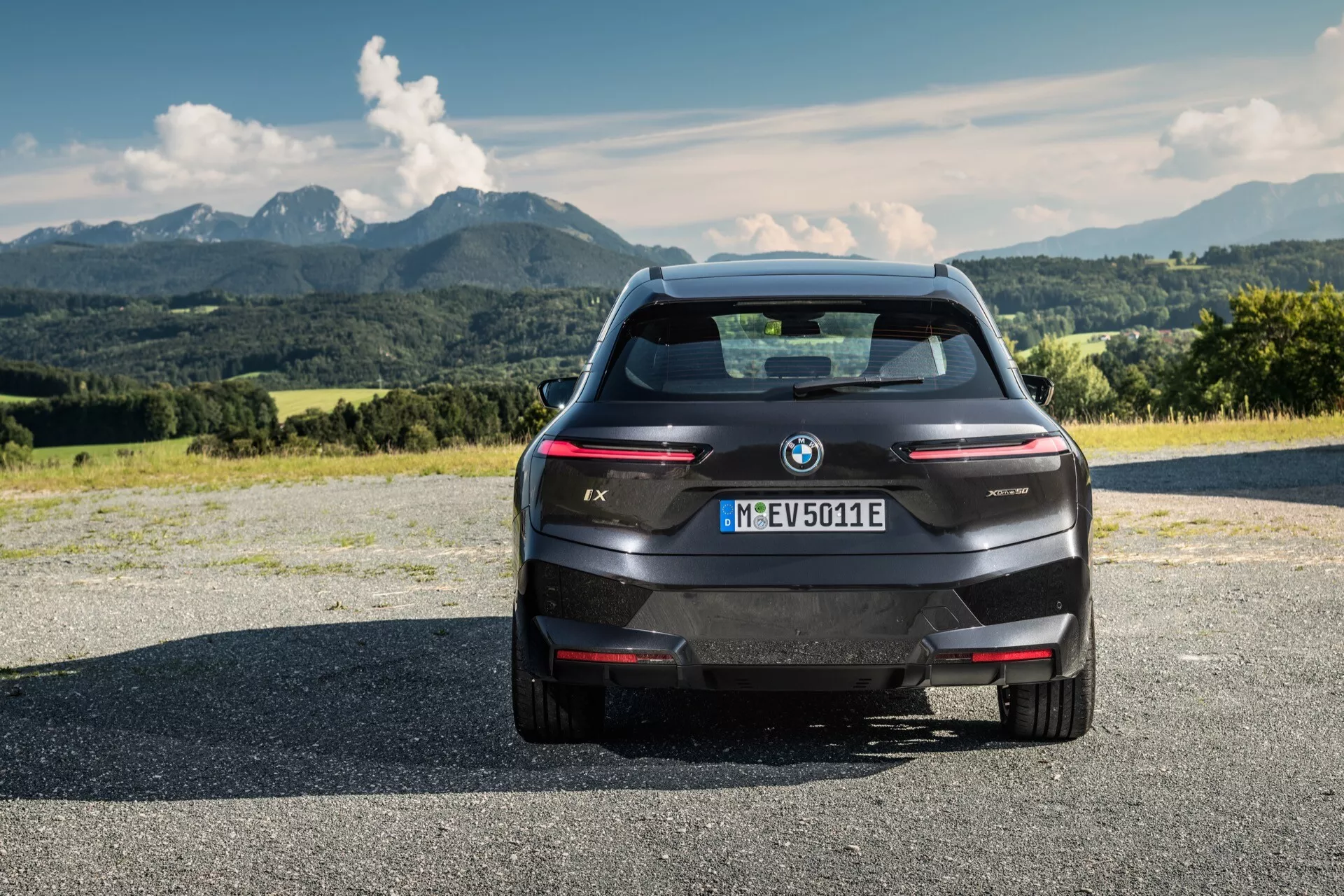 Vista trasera de un SUV eléctrico BMW iX 2021 sobre una superficie de asfalto con montañas y un cielo parcialmente nublado en el fondo, mostrando su diseño moderno, líneas elegantes, logotipo de BMW y la designación del modelo, sensores de estacionamiento traseros integrados, luz de freno montada en alto, cámara trasera, luces traseras delgadas horizontales y una ventana trasera aerodinámica sin un escape tradicional, indicando su naturaleza eléctrica y registro en Múnich.
