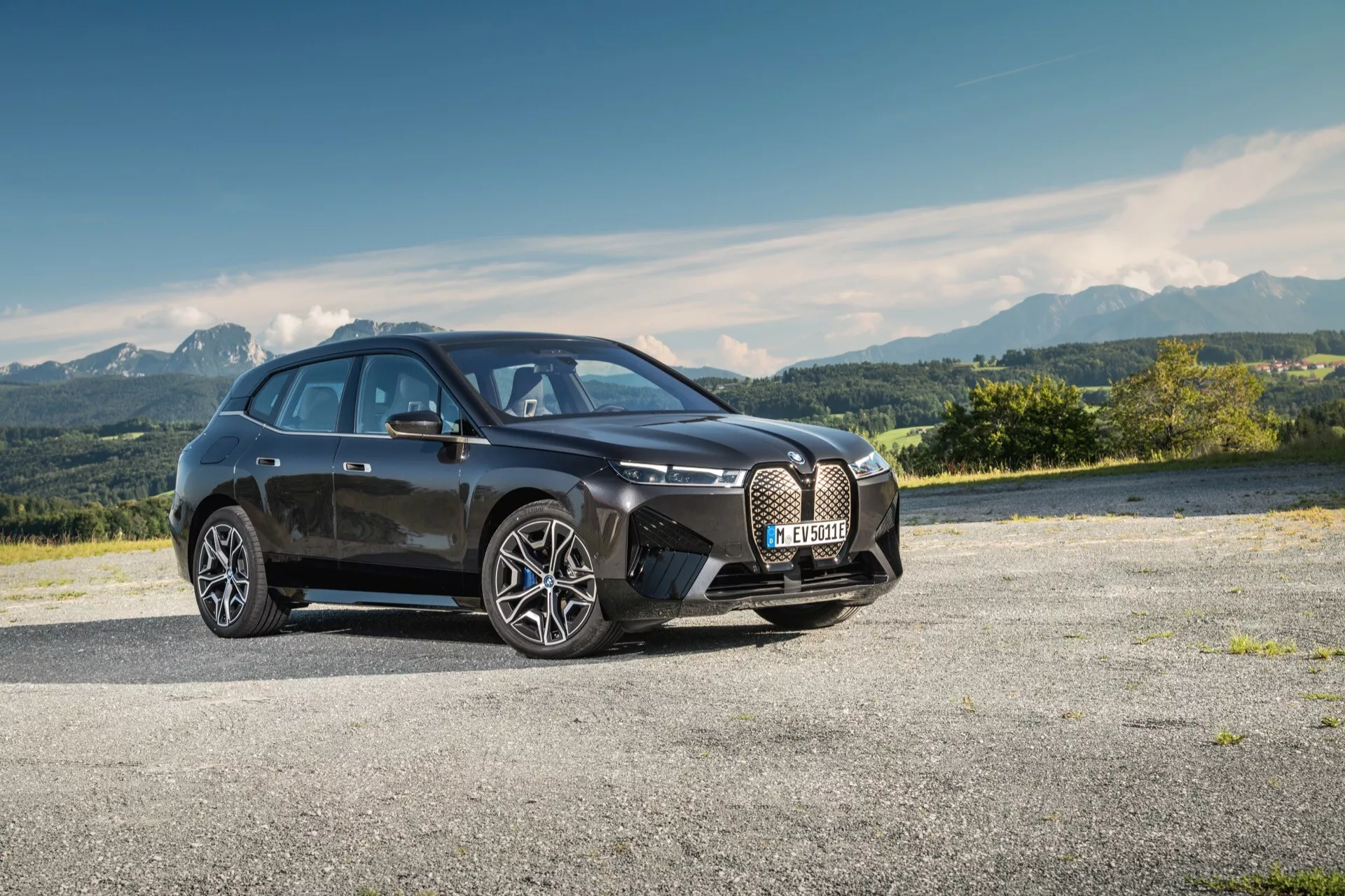 Hatchback moderno de cuatro puertas BMW iX (i20) de 2021 estacionado en un área pavimentada con paisaje montañoso al fondo, mostrando su gran parrilla en forma de riñón, líneas elegantes, faros LED y diseño aerodinámico con grandes ruedas de aleación