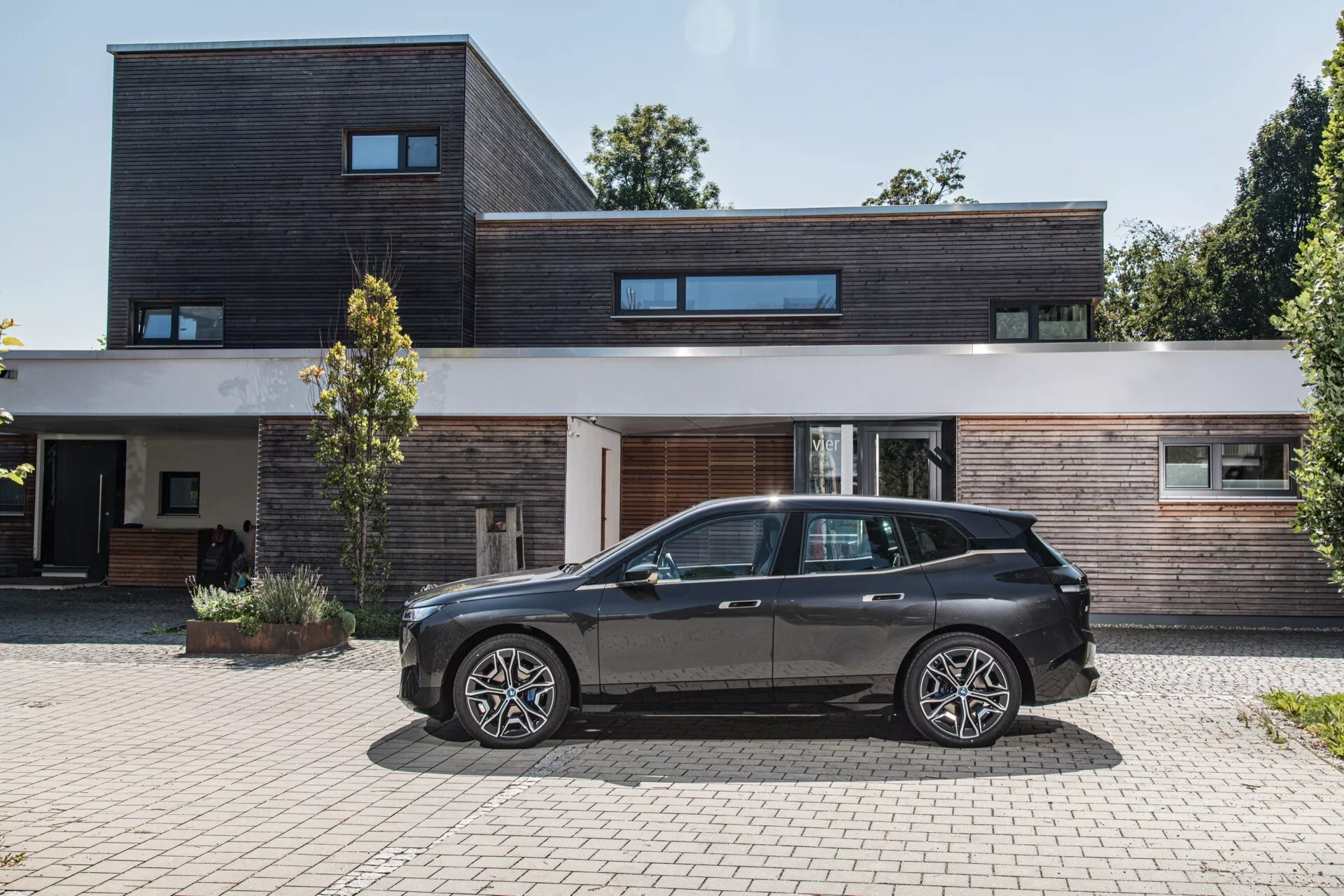 BMW iX (i20) del 2021 estacionado frente a una casa moderna con revestimiento de madera y materiales oscuros, mostrando el perfil lateral del coche, diseño aerodinámico, ruedas de aleación y alta altura del suelo