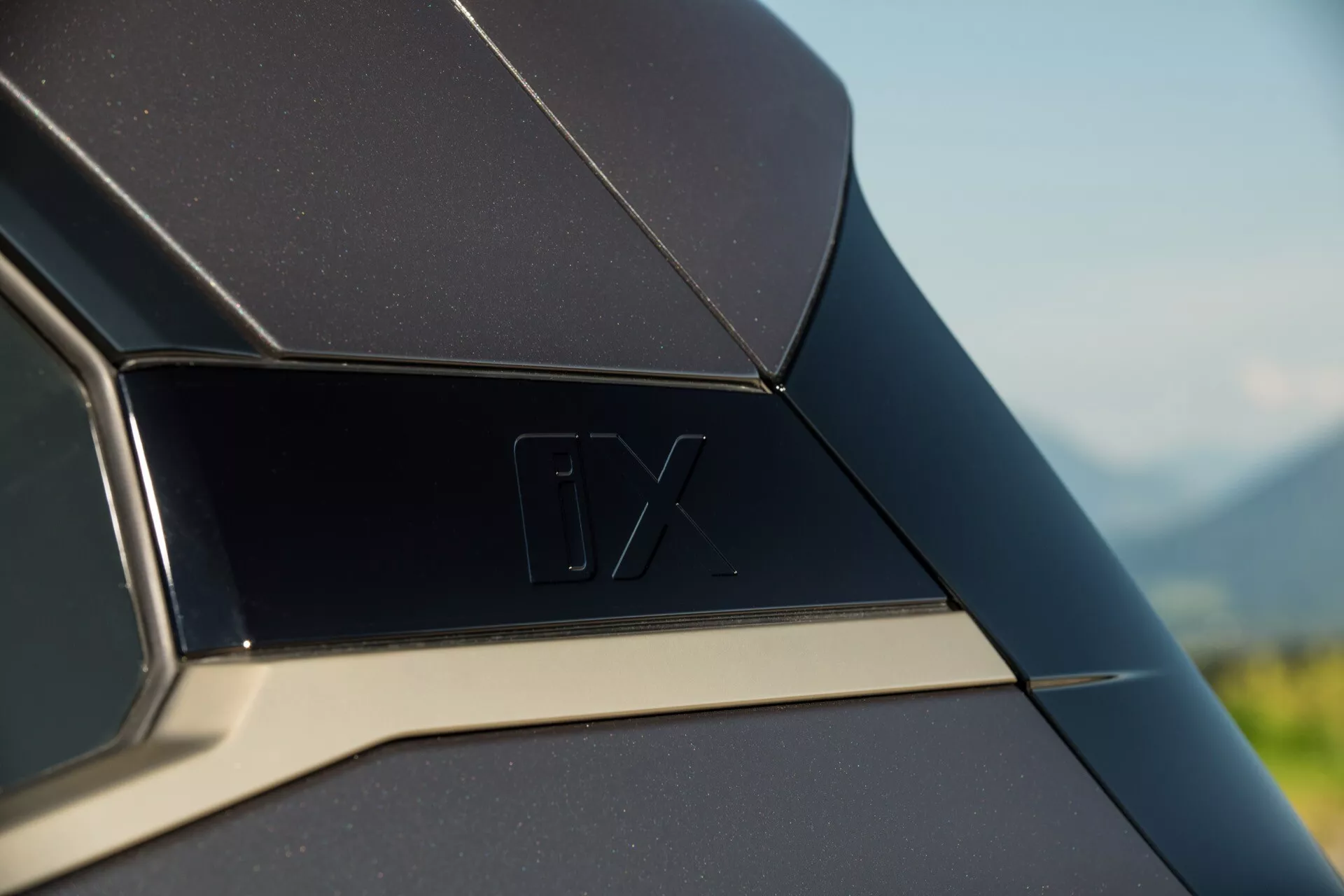 Imagen detallada del detalle exterior del BMW iX 2021, mostrando el panel trasero o el área del pilar C con el distintivo 'iX' y una línea de adorno cromado contrastante, contra un fondo natural desenfocado