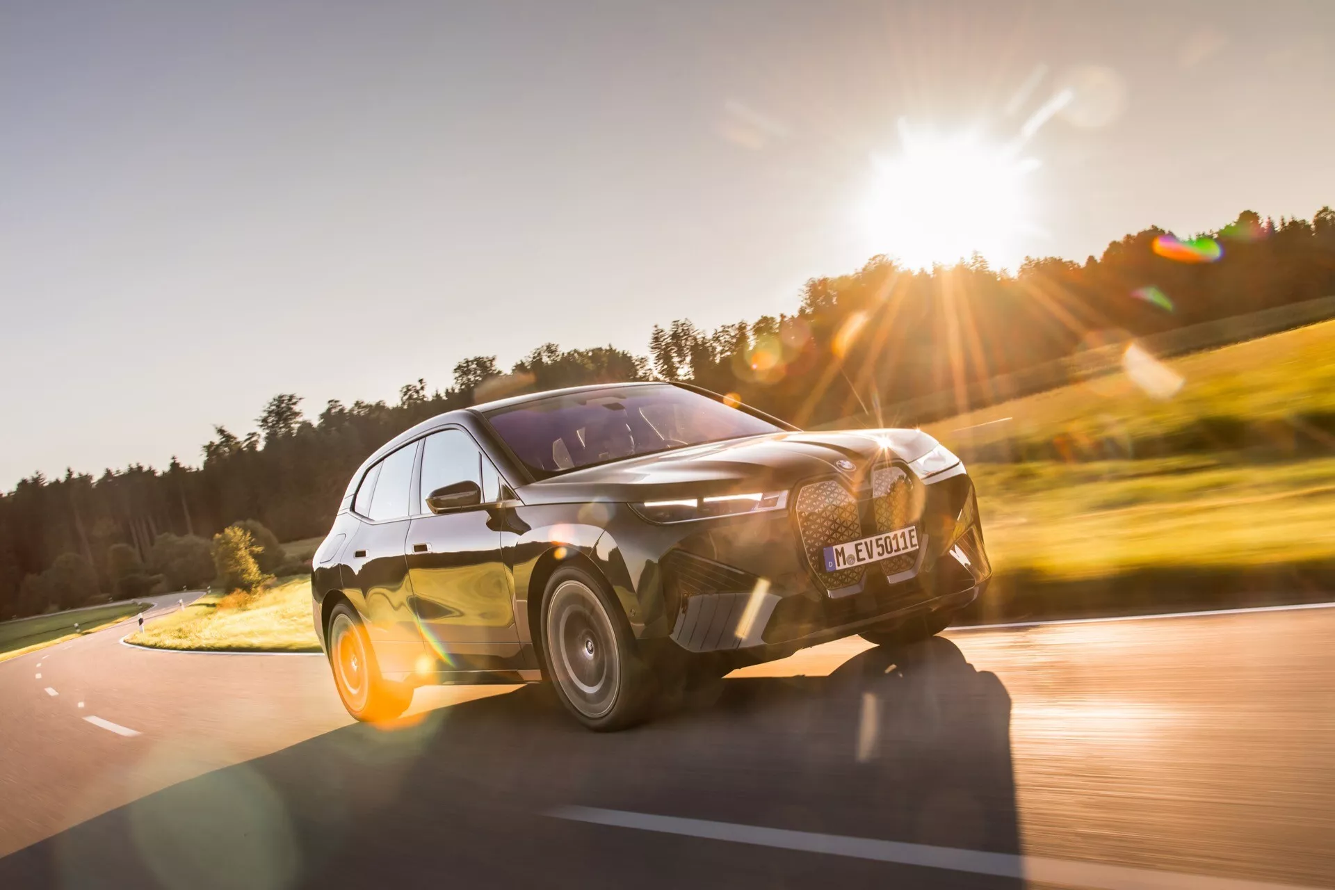 Vista frontal dinámica de tres cuartos del BMW iX 2021 en movimiento, destacando su postura deportiva, faros delanteros LED elegantes y la distintiva parrilla grande, capturada desde un ángulo bajo para enfatizar las características de diseño moderno y agresivo del automóvil contra un telón de fondo rural natural.