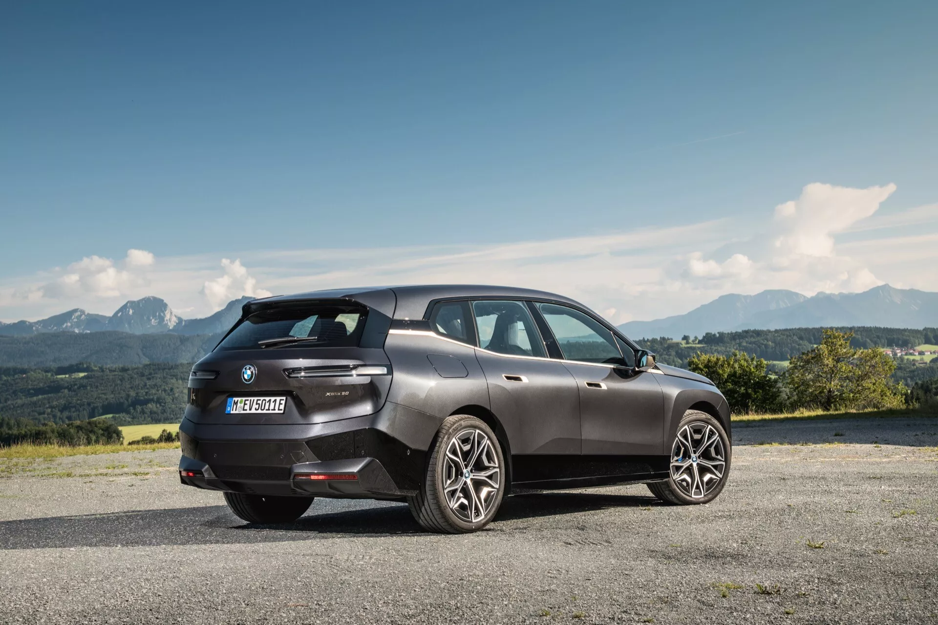 Vista trasera de tres cuartos de un BMW iX 2021 en gris oscuro, aparcado en un pavimento elevado con un paisaje montañoso al fondo, reflejando un diseño moderno de SUV de lujo