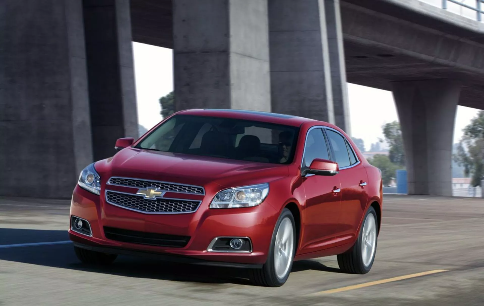 Vista dinámica de un sedán Chevrolet Malibu 2012 rojo en movimiento sobre un puente, mostrando el diseño frontal con parrilla cromada, faros elegantes, silueta deportiva y ruedas de aleación.