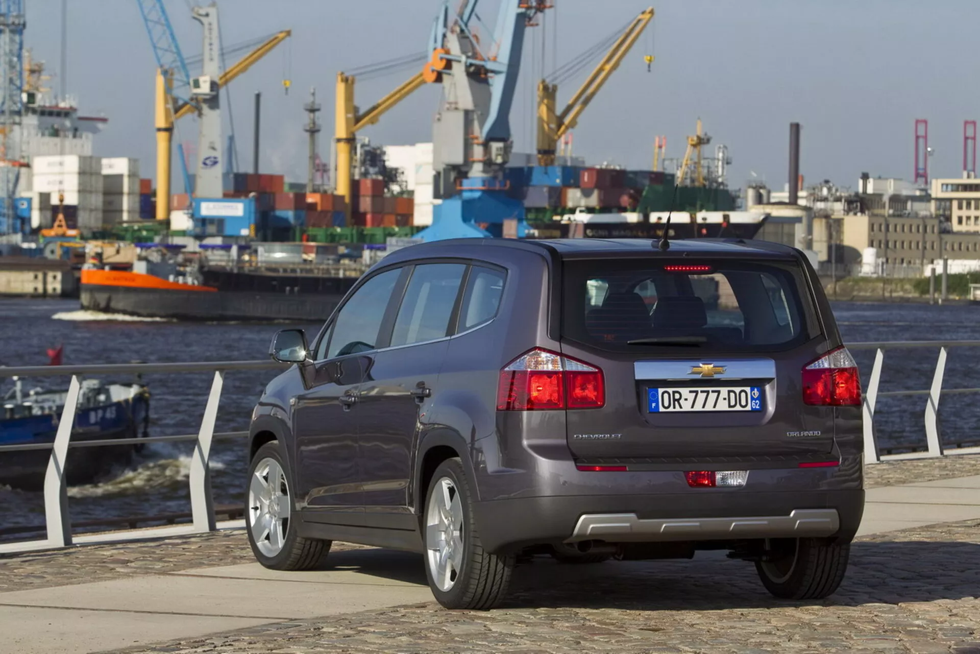 Vista trasera de tres cuartos del Chevrolet Orlando 2011 en un muelle frente al agua, destacando su carrocería de SUV compacto, configuración de cinco puertas, sistema de escape doble, sensores de asistencia de estacionamiento en el parachoques y ruedas de aleación de múltiples radios