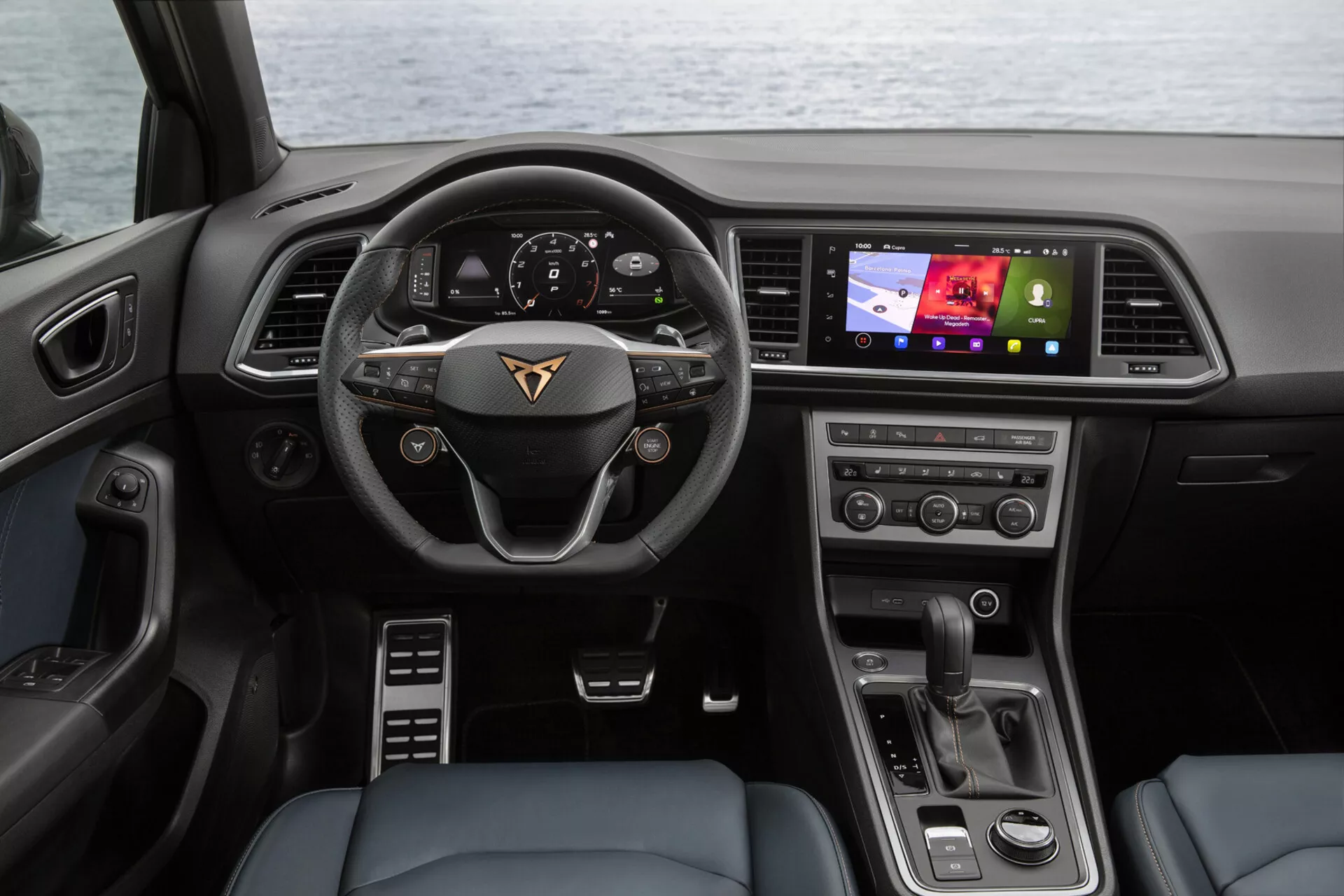 Vista interior del Cupra Ateca 2020 desde el asiento trasero mostrando el tablero de instrumentos, volante multifuncional con emblema del fabricante, cuadro de instrumentos digital, pantalla táctil de infoentretenimiento orientada al conductor, botones de control climático y selector de marchas moderno
