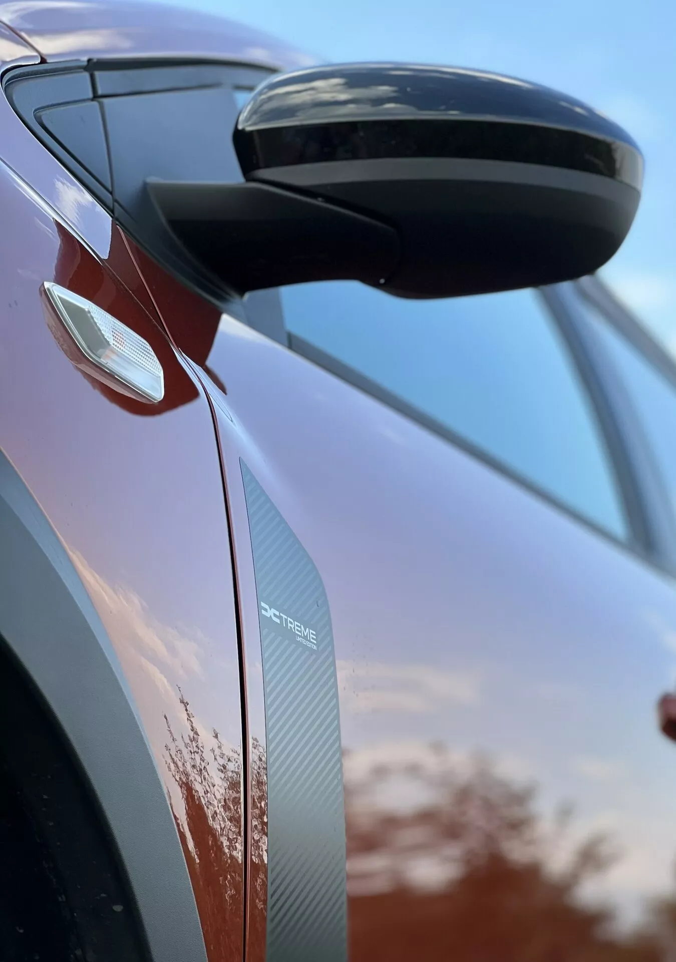 Detalle cercano del Dacia Jogger 2022 centrado en el espejo retrovisor lateral y el diseño de la puerta, mostrando características aerodinámicas, señales de giro integradas y texturas de fibra de carbono contra un fondo de cielo