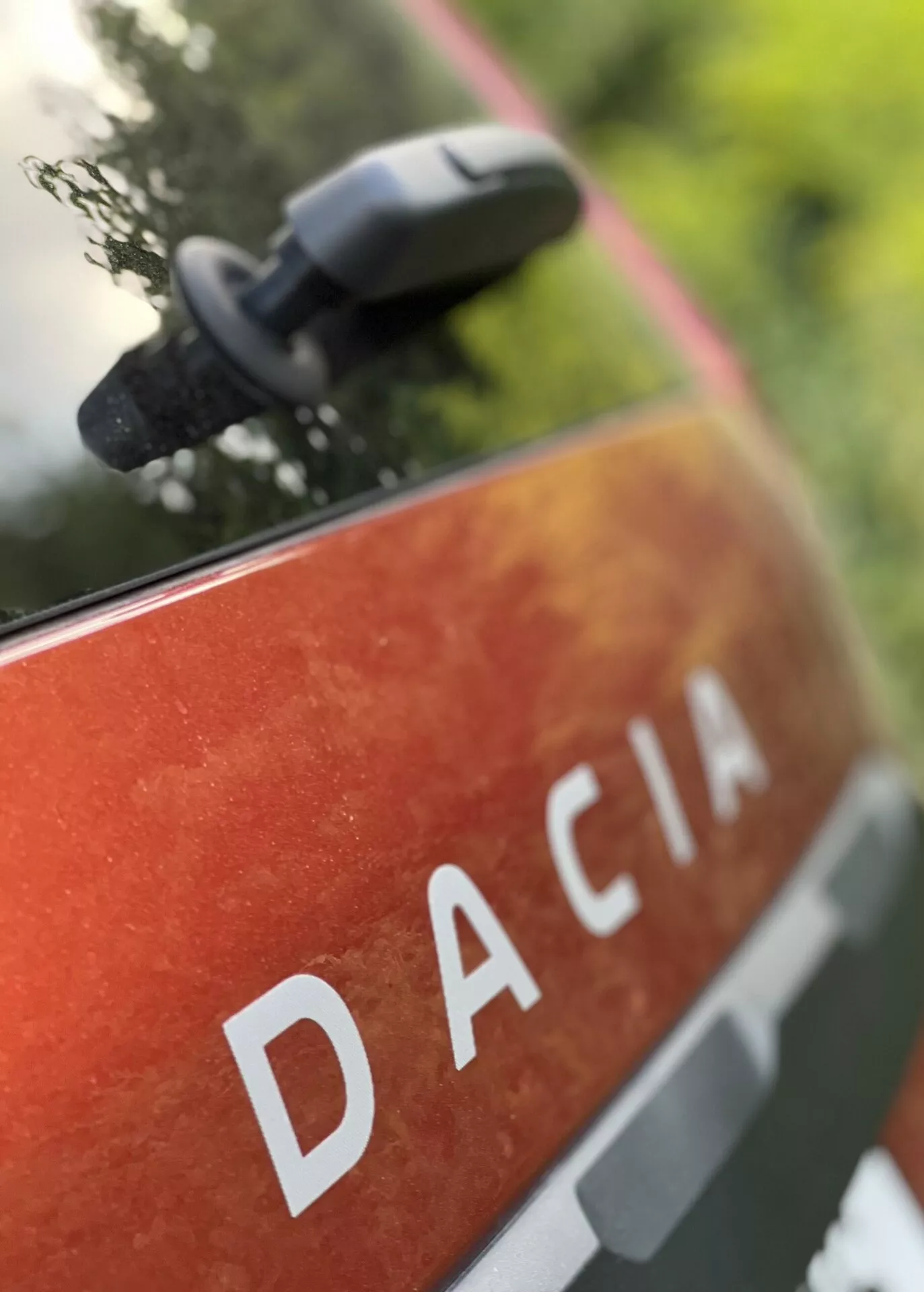 Vista detallada de la parte trasera del Dacia Jogger 2022 con el nombre DACIA en letras mayúsculas blancas sobre una pintura metalizada naranja-roja con motas y suciedad, sugiriendo uso y durabilidad al aire libre, situado en un entorno natural con un espejo lateral desenfocado que indica características modernas del espejo.