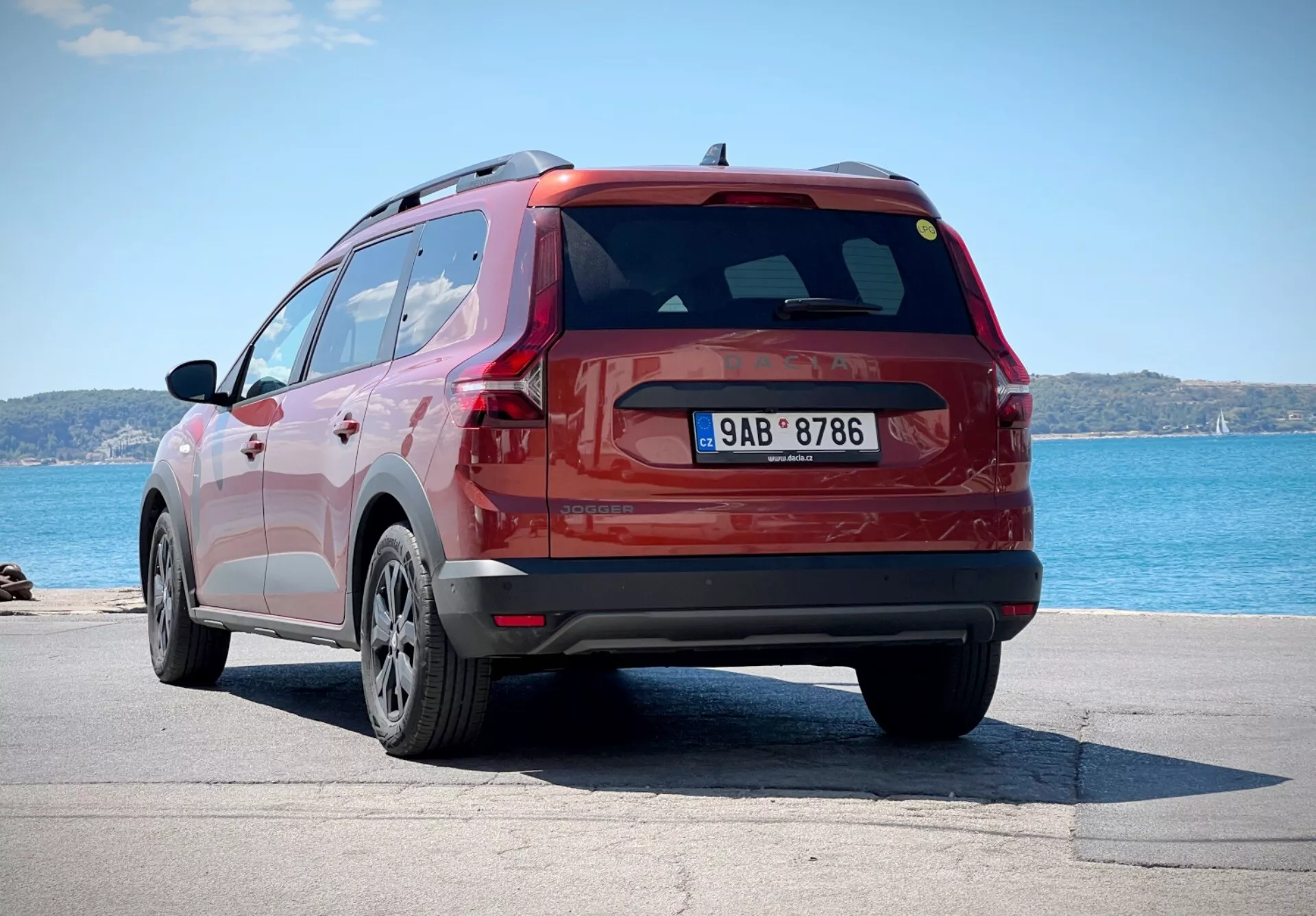 Dacia Jogger 2022 en rojo, capturado desde un ángulo trasero de tres cuartos que muestra el costado del SUV y el diseño del portón trasero, aparcado cerca de un cuerpo de agua con el horizonte en el fondo, destacando sus ruedas de aleación de múltiples radios, luces traseras LED y alerón trasero aerodinámico